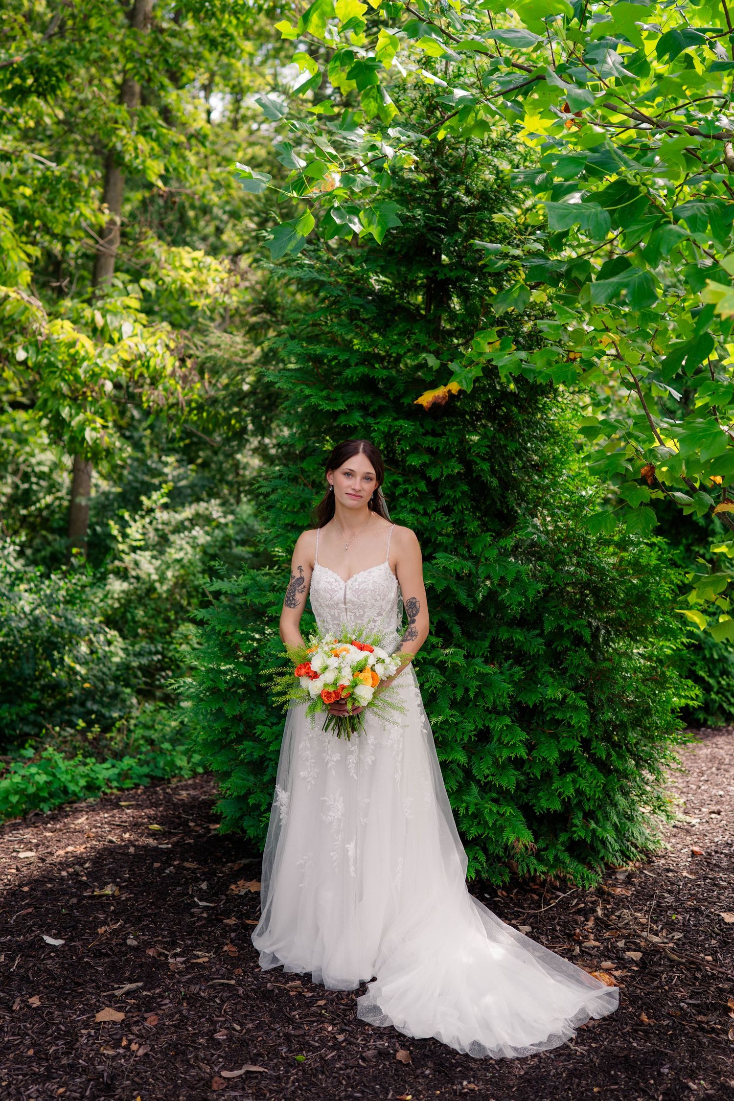 Bride in lace wedding gown holds colorful bouquet while posing in lush green garden setting.