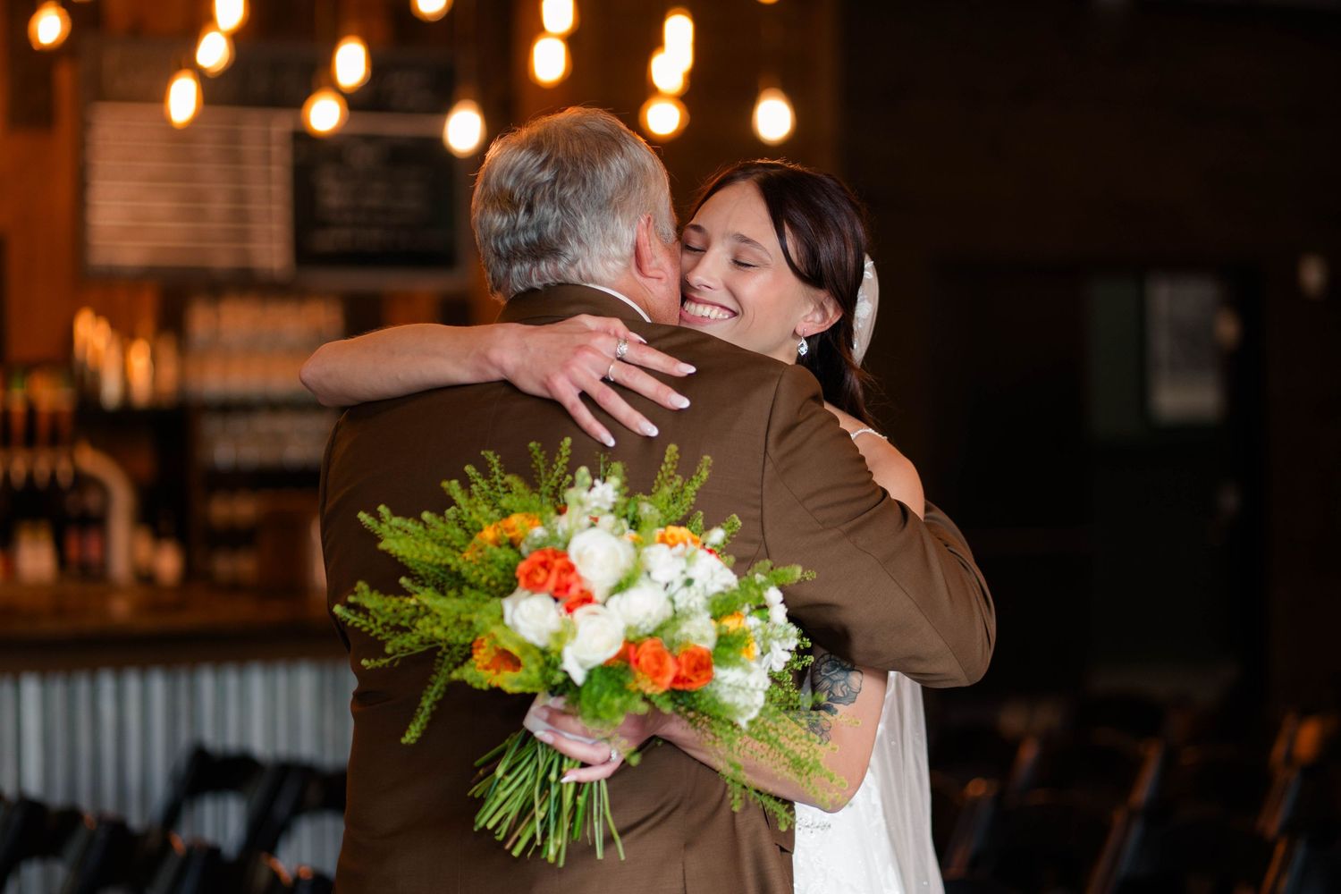 Bride holding colorful bouquet embracing guest in rustic venue with warm lighting.