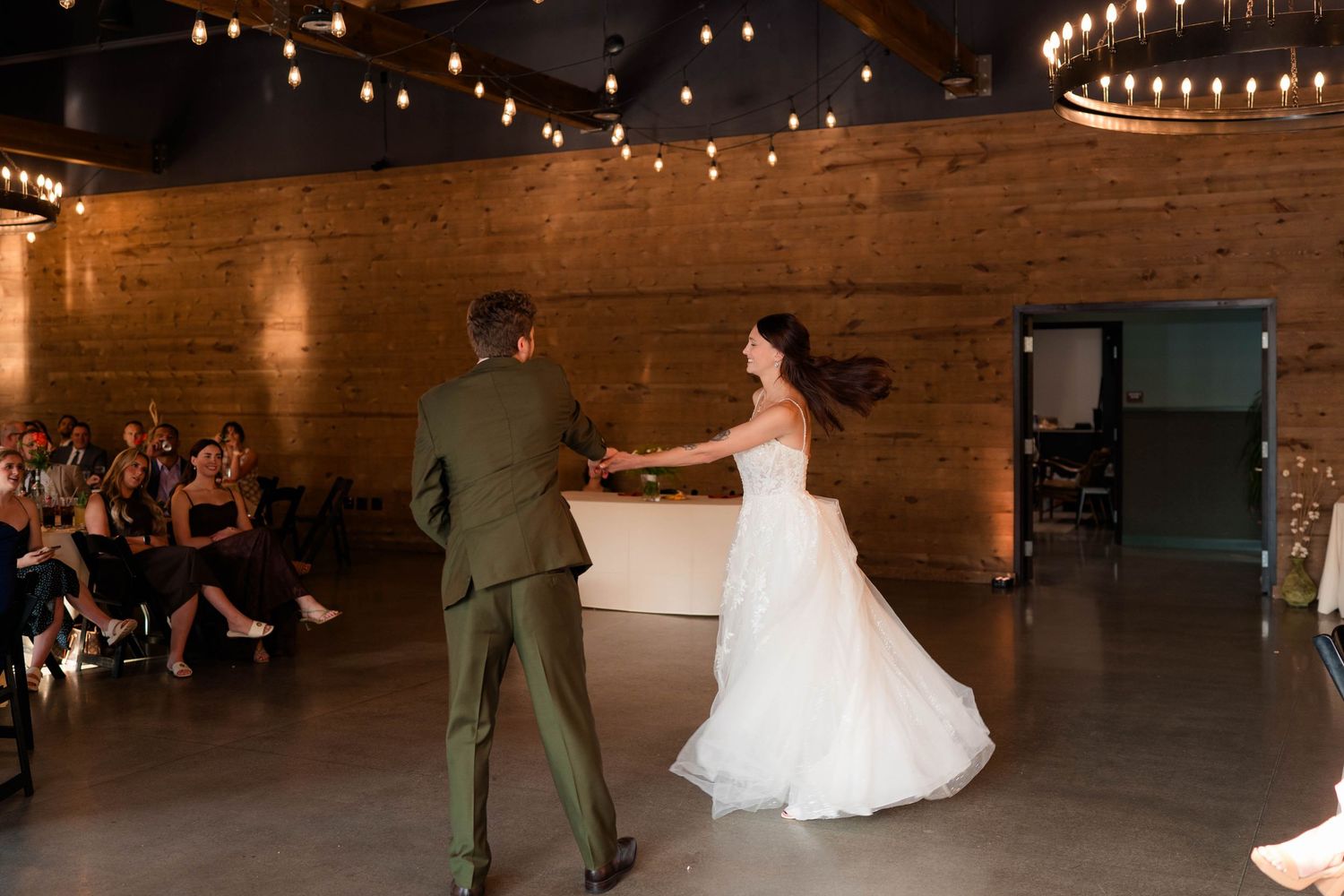 Bride in white gown spins during first dance with groom in olive suit in a warmly lit reception venue.