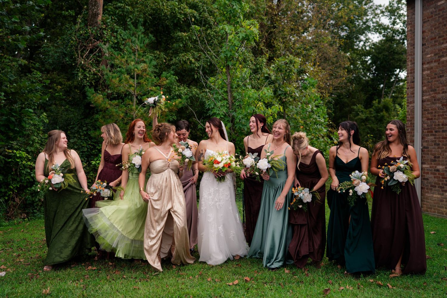 Bride laughs joyfully with bridesmaids in mismatched earth-tone gowns on a lush green outdoor setting.