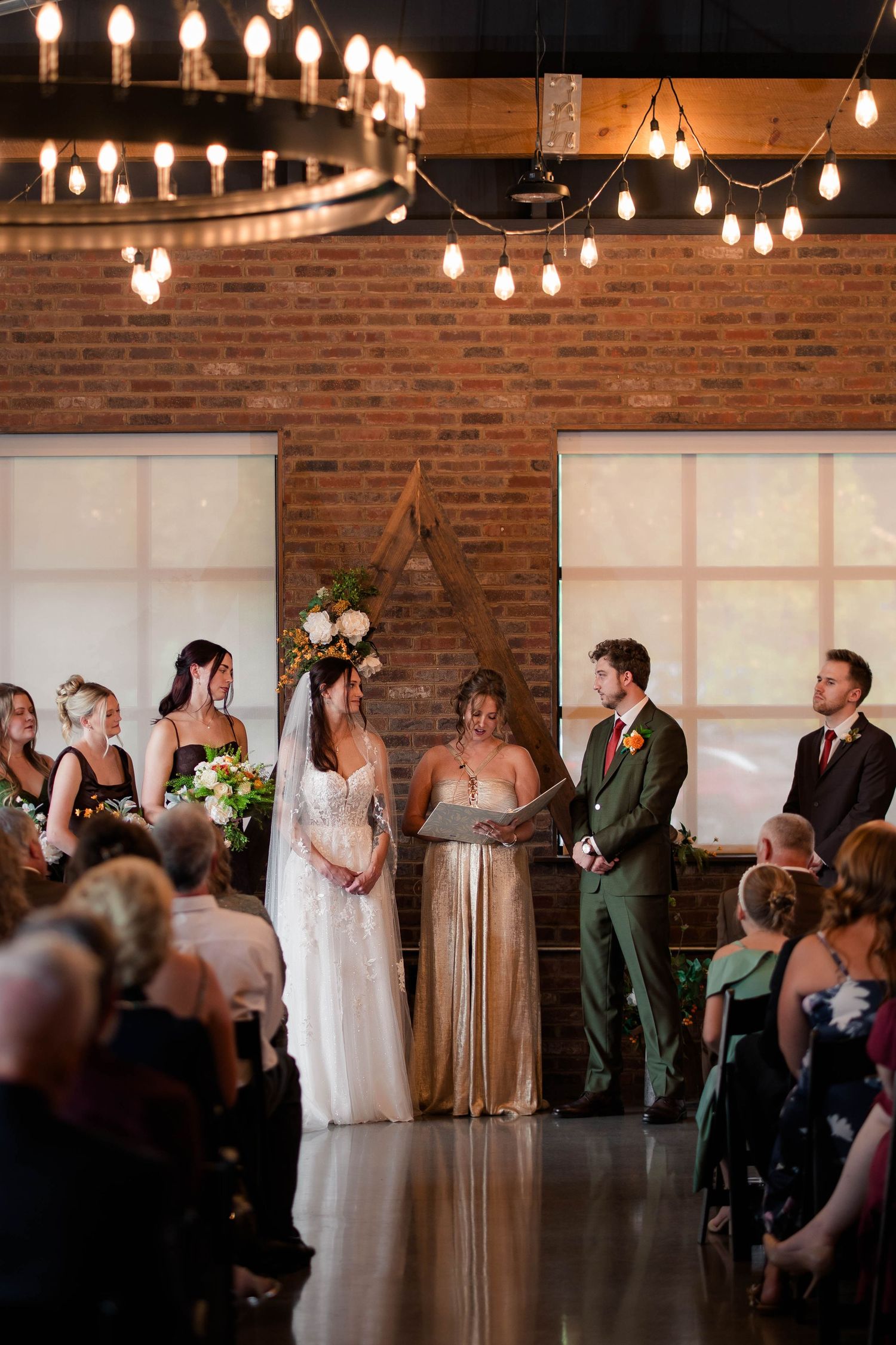Bride and groom exchange vows under Edison bulb chandeliers in rustic brick wedding ceremony venue.
