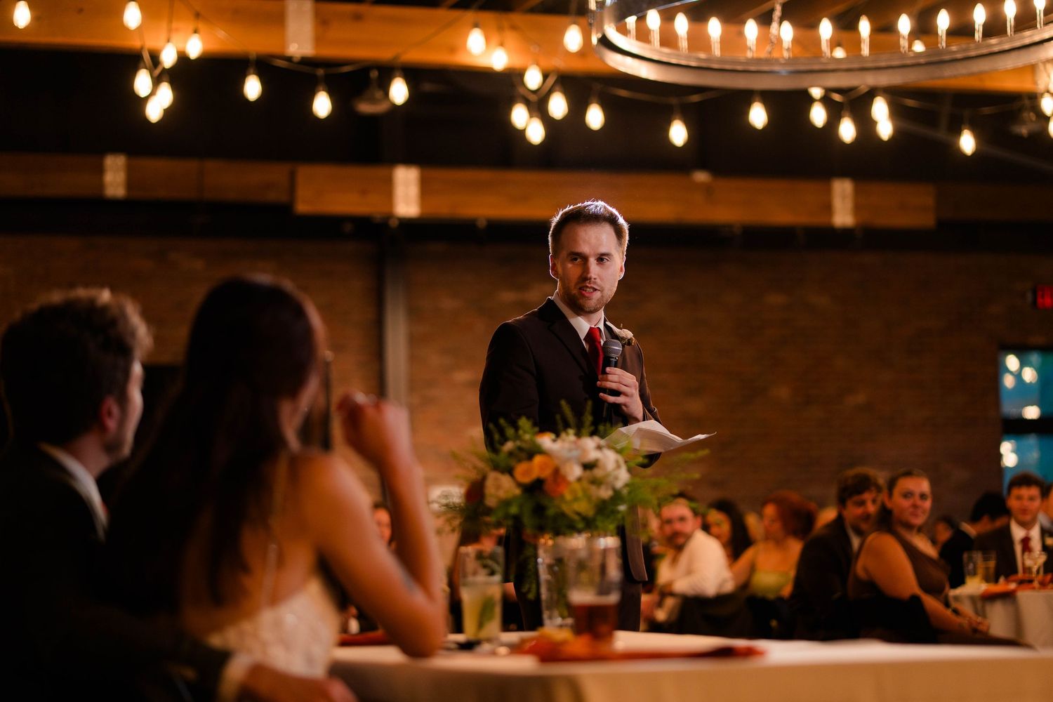 Man in dark suit delivers wedding toast holding paper while guests listen at reception.