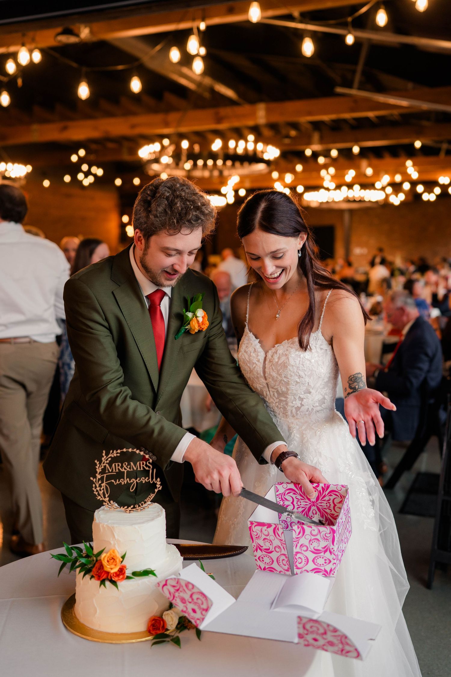 Bride and groom in green suit cutting white wedding cake under warm string lights at reception.