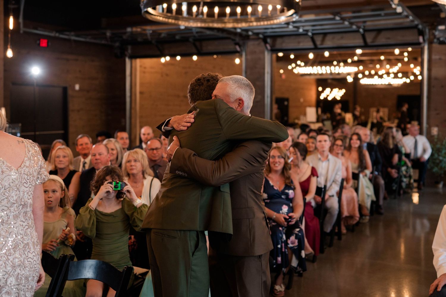 Emotional groom embracing father during wedding ceremony processional at rustic indoor venue.