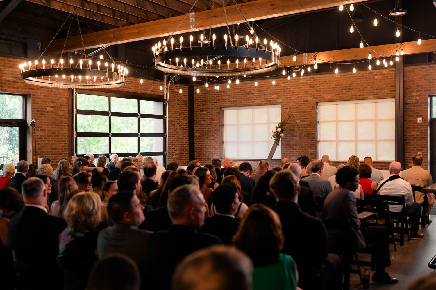 Guests seated at elegant indoor wedding ceremony in rustic brick venue with chandelier string lights.