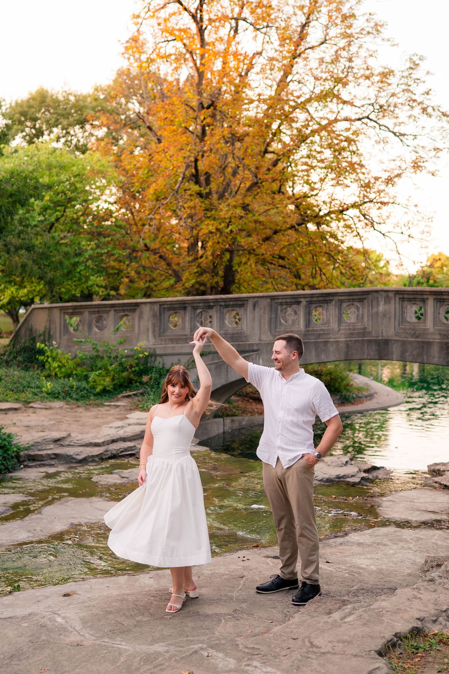 Man twirls woman in white dress on rocks by a pond with a stone bridge and autumn tree.