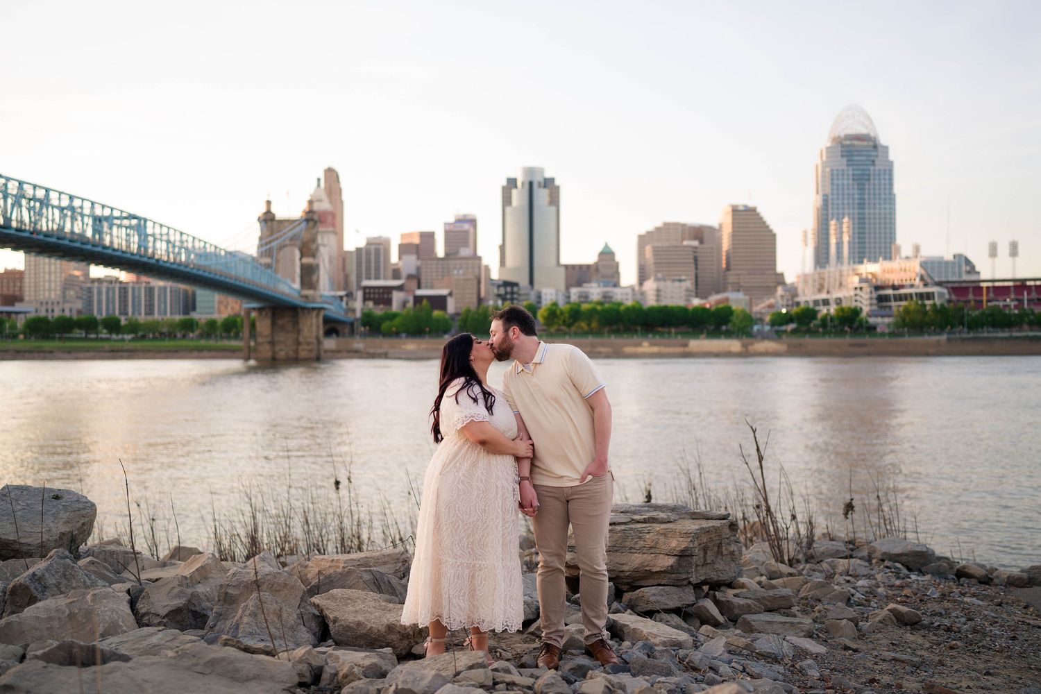 Couple sharing a romantic kiss by the river with the Cincinnati city skyline and bridge at sunset.