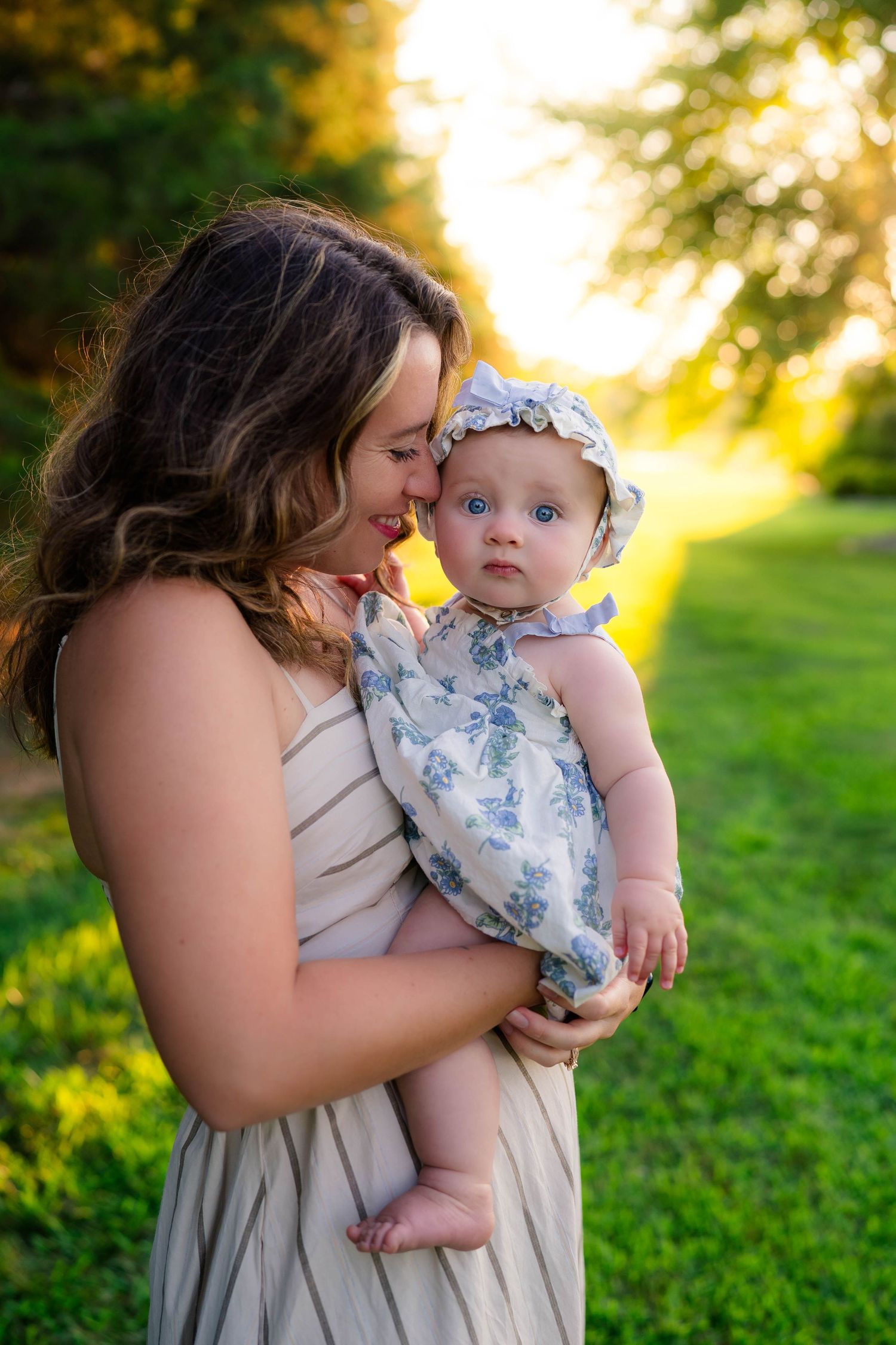 A young mother nuzzles her wide-eyed baby girl in a floral outfit during a golden sunset outdoors.