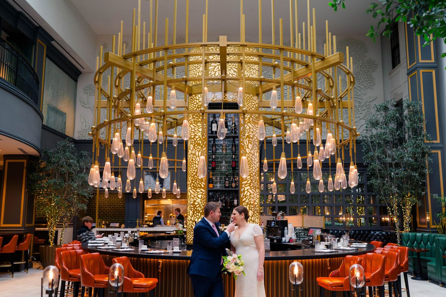 Bride and groom posing beneath a stunning circular gold chandelier in an upscale wedding venue bar.