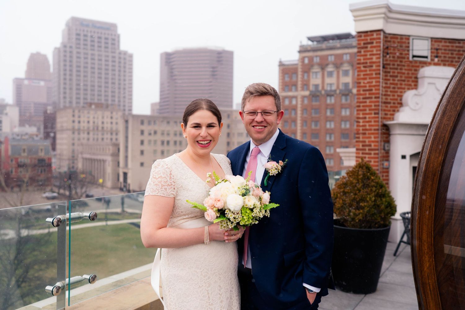 Smiling bride and groom posing on rooftop terrace with city skyline backdrop on their wedding day.