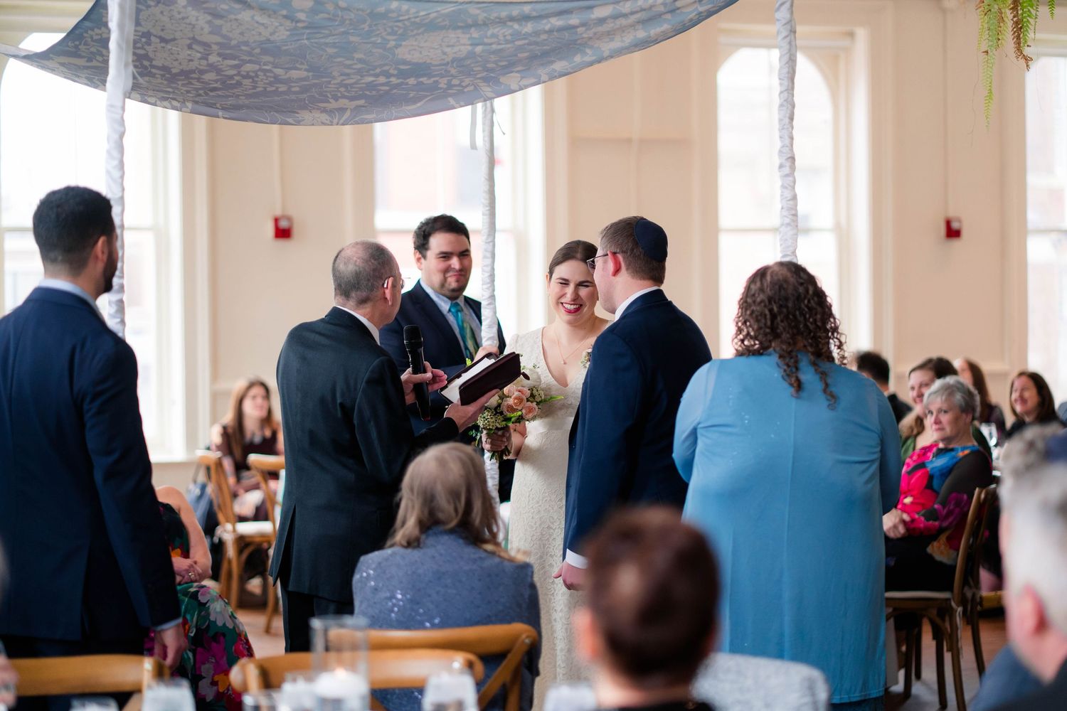 Wedding party stands under a chuppah during an indoor Jewish ceremony with seated guests.