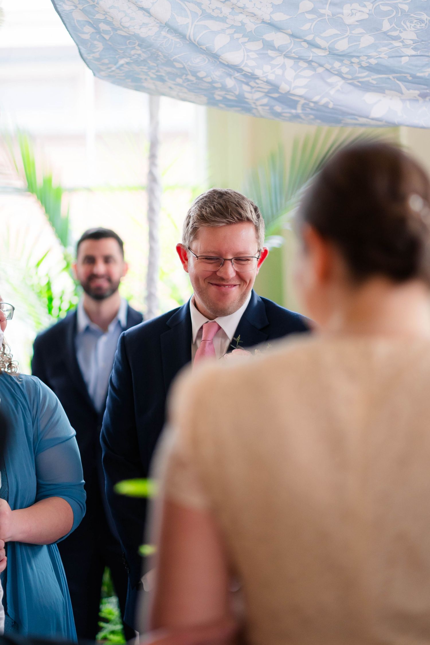 Groom smiles warmly at bride during indoor wedding ceremony with lush tropical greenery backdrop.