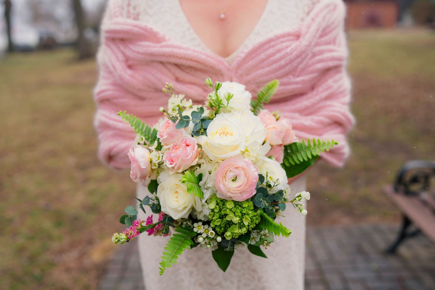 Close-up of a bridal bouquet with white and pink ranunculus, roses, and greenery held by bride in pink shawl.