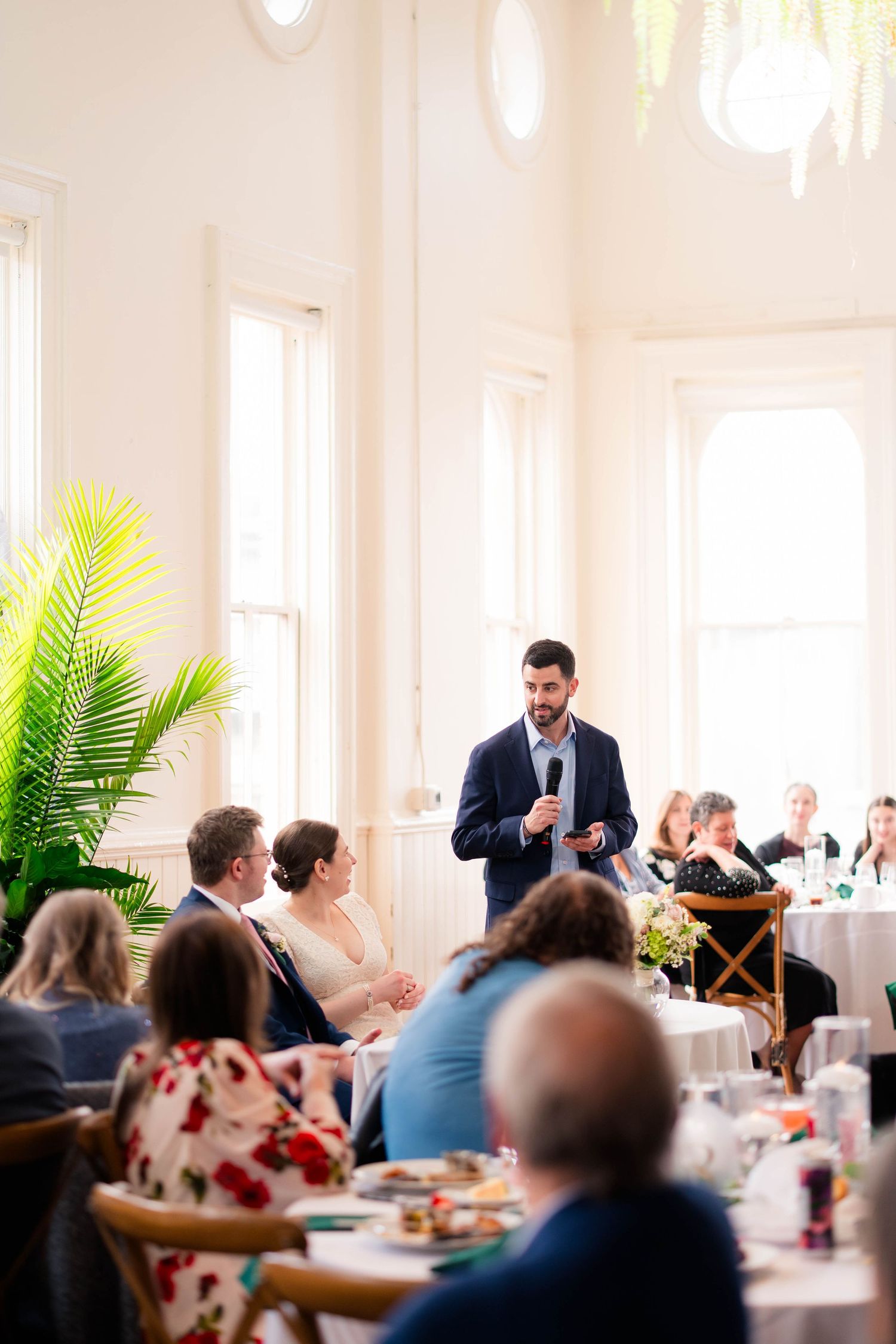 Wedding speaker delivers toast in bright elegant venue while guests and couple listen attentively.