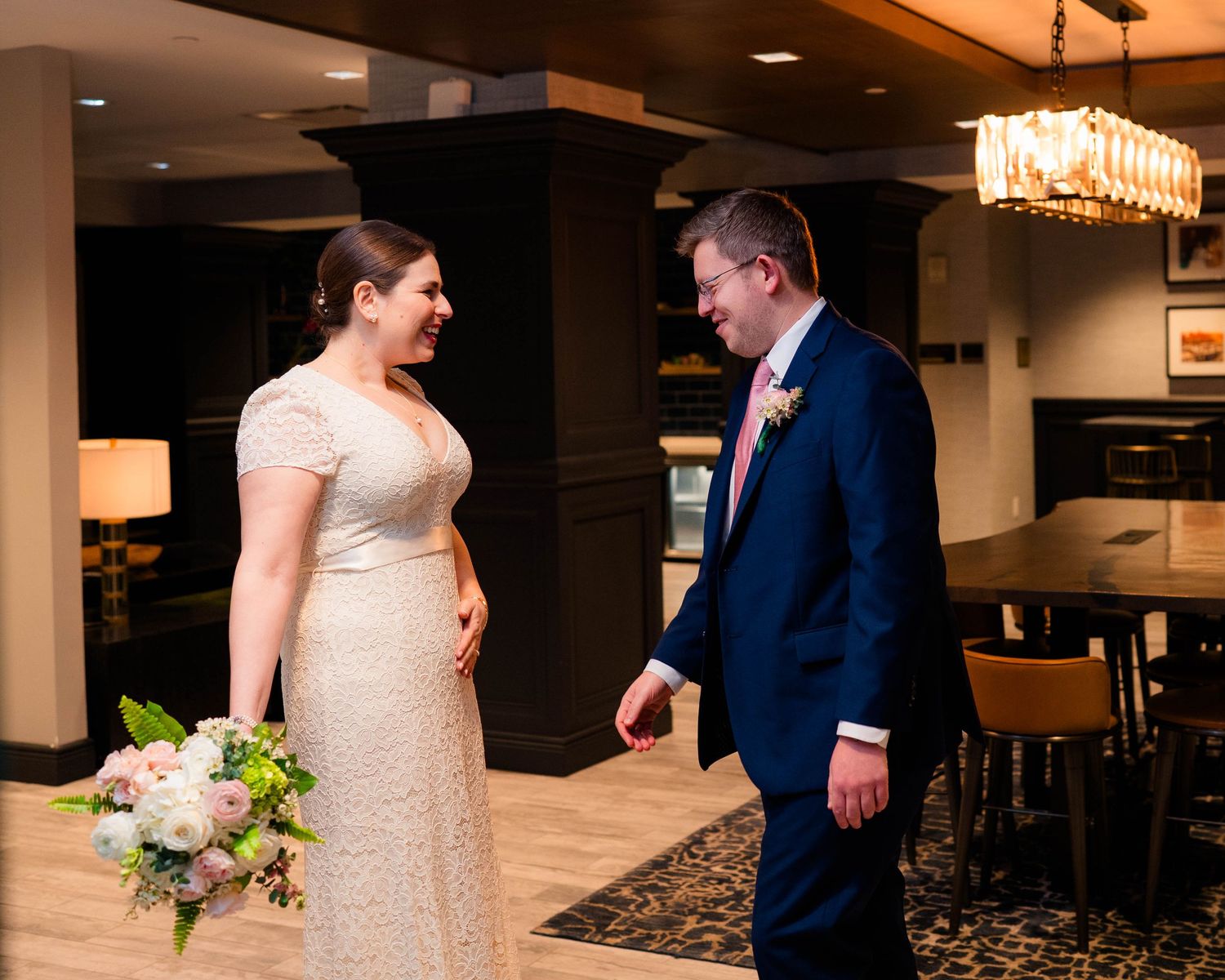 Bride in lace gown holding bouquet meets groom in navy suit for first look in hotel lobby.