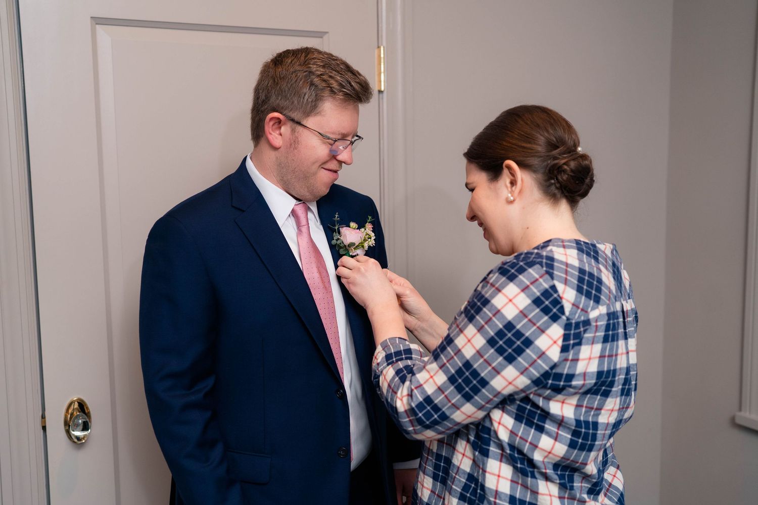 Woman in plaid shirt pinning a floral boutonniere on a groom in a navy suit.