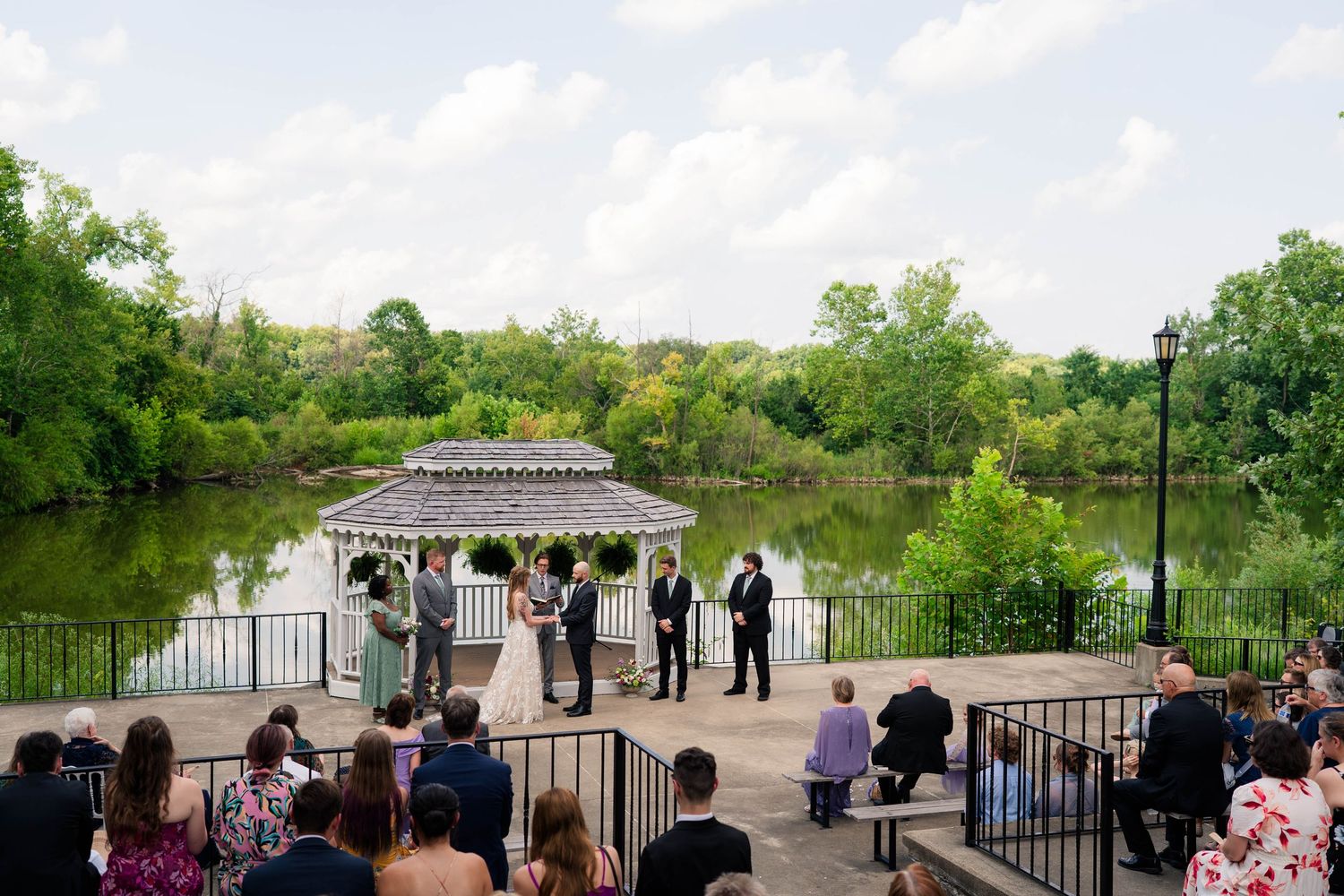 Outdoor wedding ceremony at a gazebo overlooking a scenic lake with guests seated.
