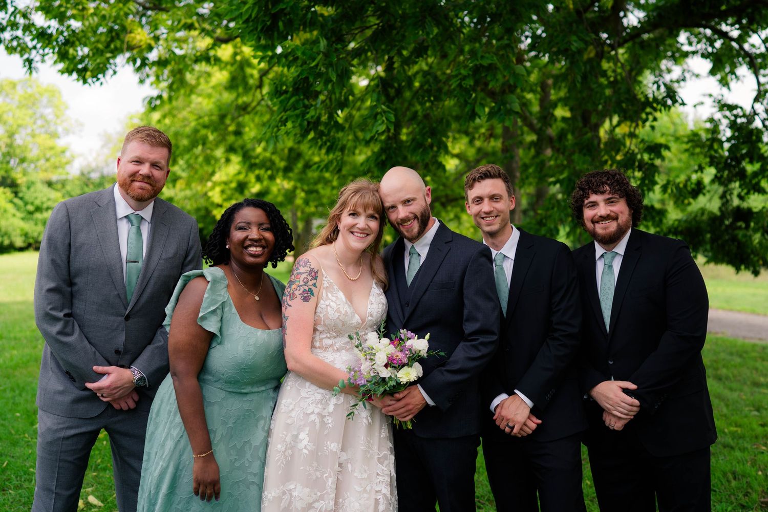 Bride and bald groom pose with four wedding party members in black and sage green attire outdoors.