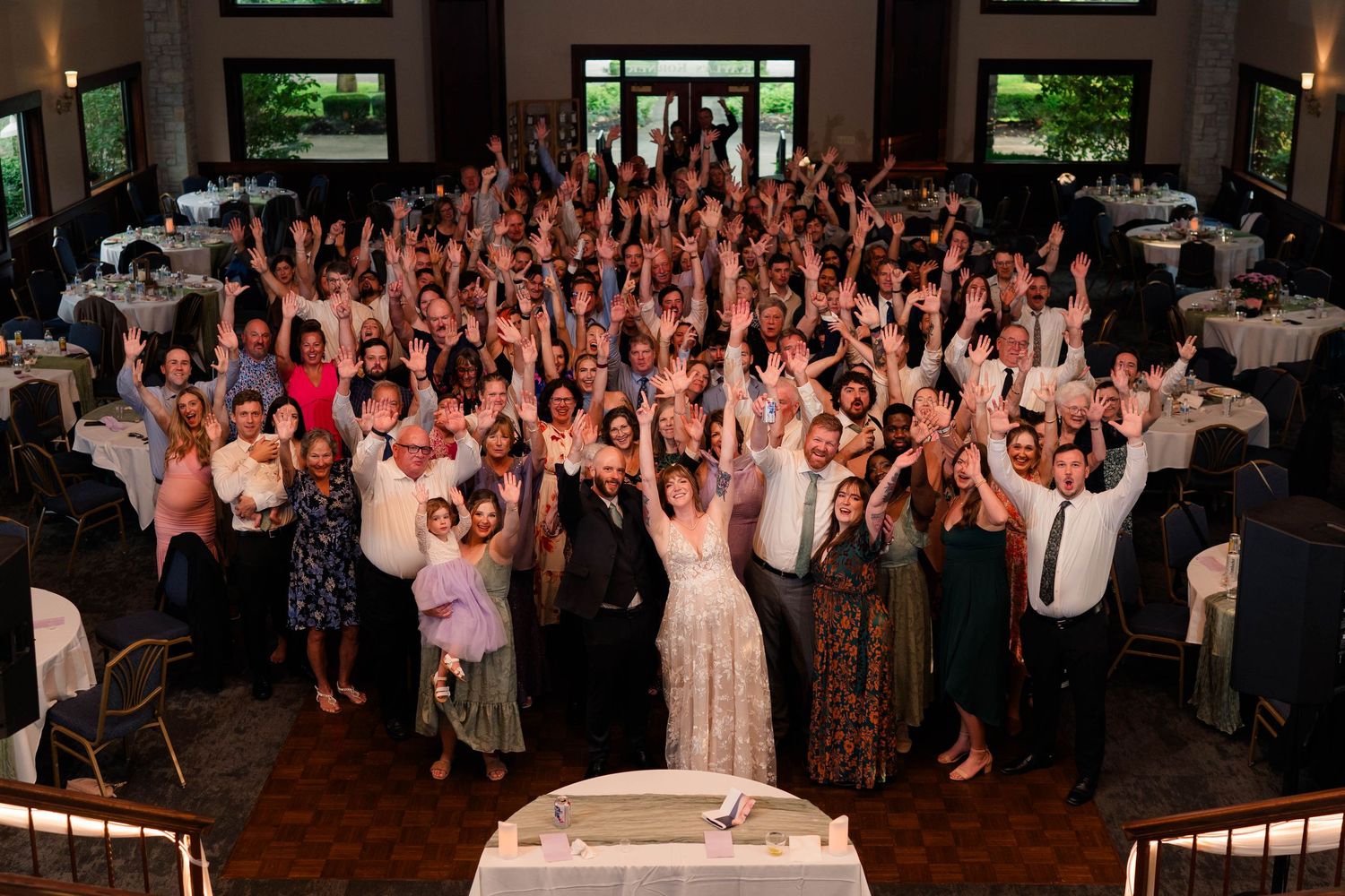 Large group of wedding guests raise hands for celebratory photo at reception venue.