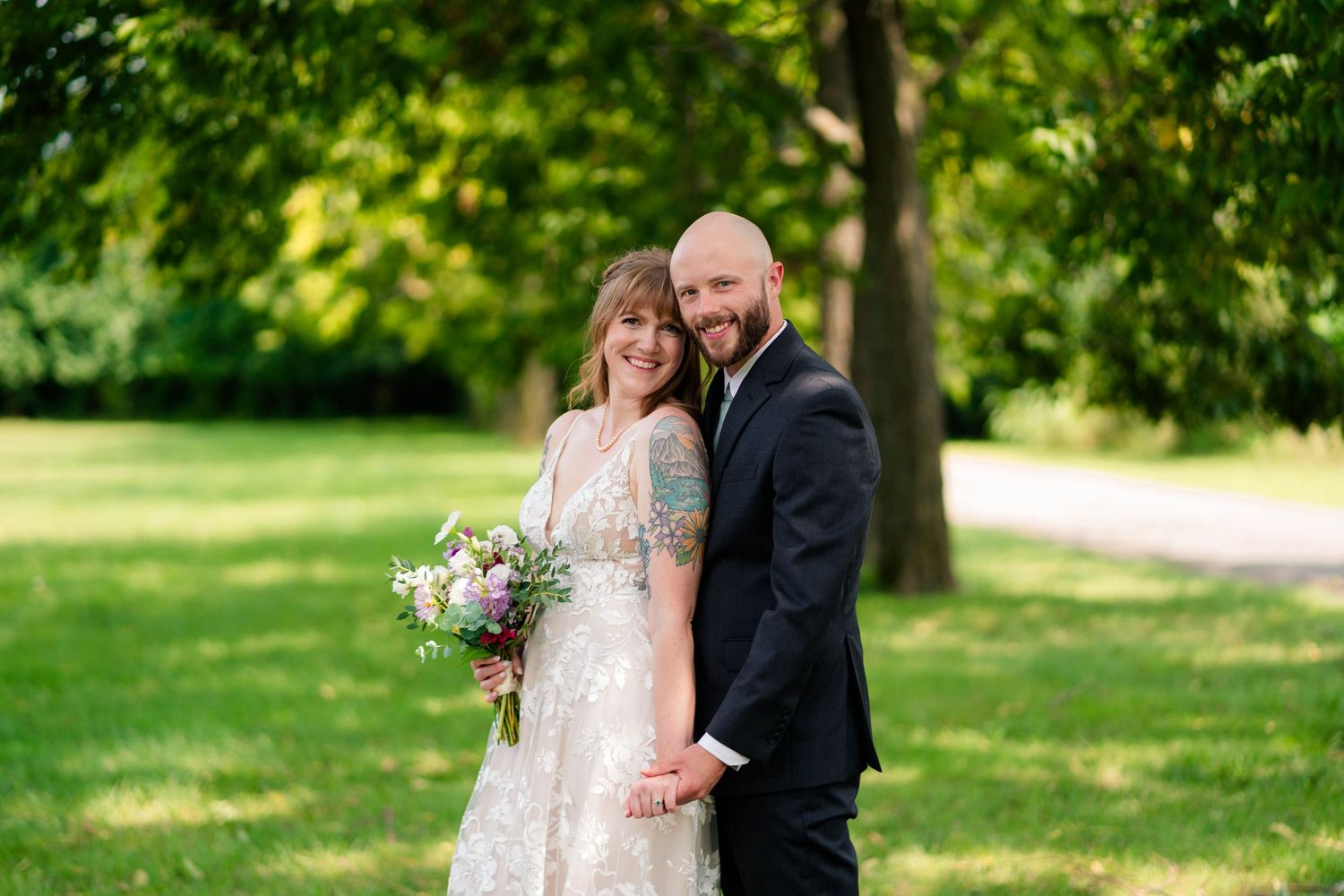 A smiling bride and groom pose together outdoors holding a wildflower bouquet on their wedding day.