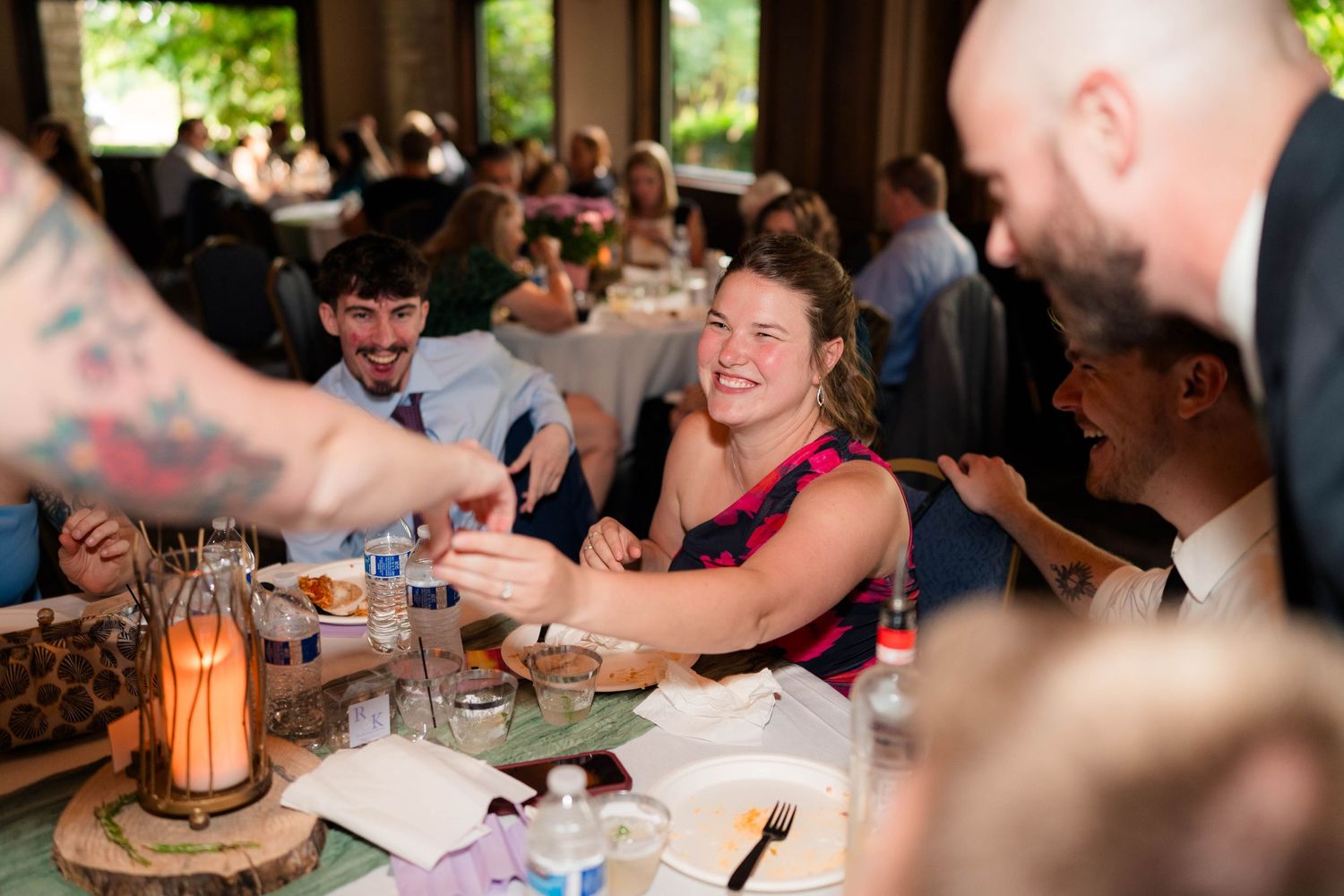 Wedding guests laugh and toast drinks at reception table decorated with rustic wooden centerpieces.