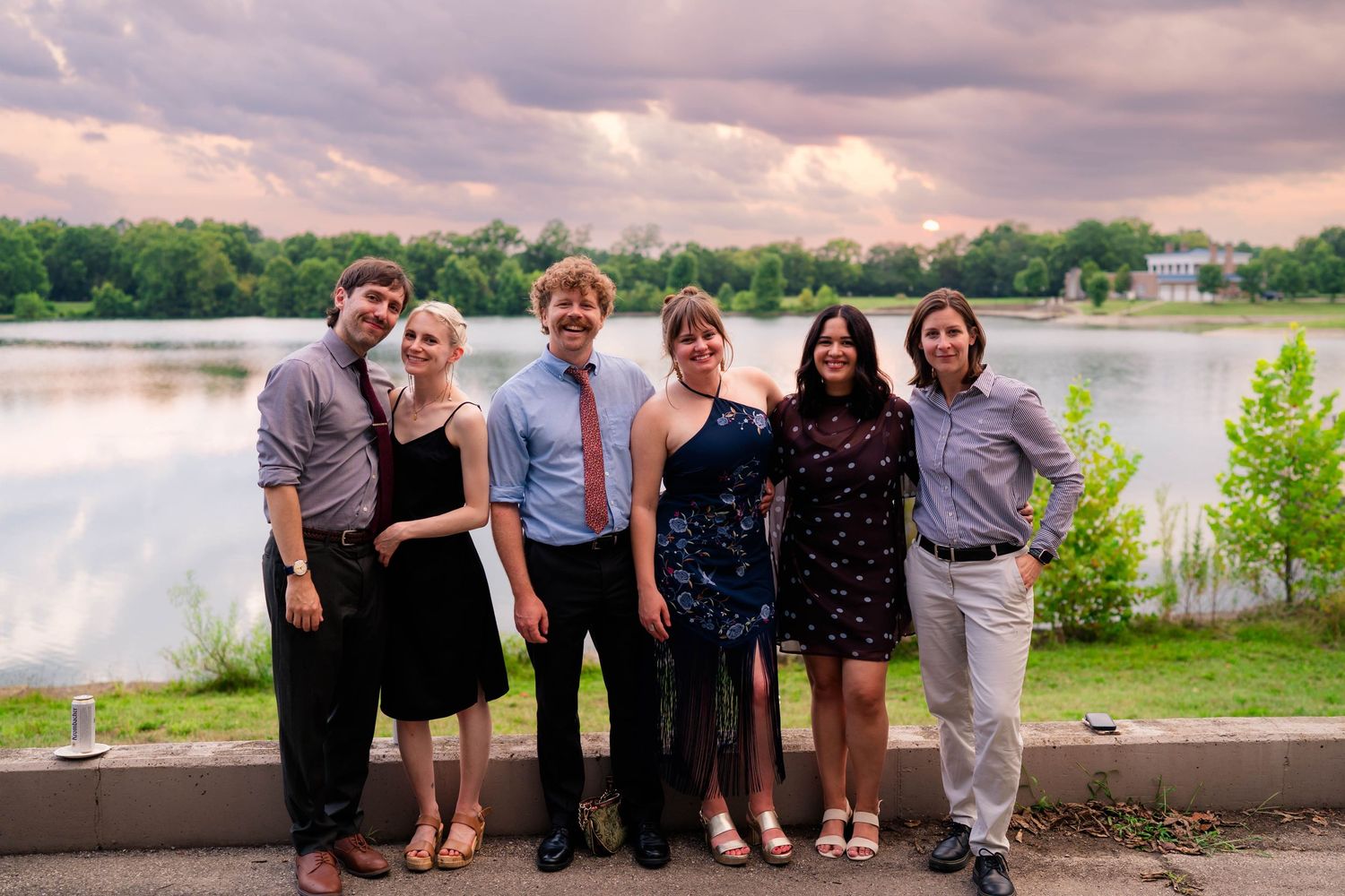 Group of six young adults pose together smiling in front of scenic lake with cloudy sunset sky behind them.