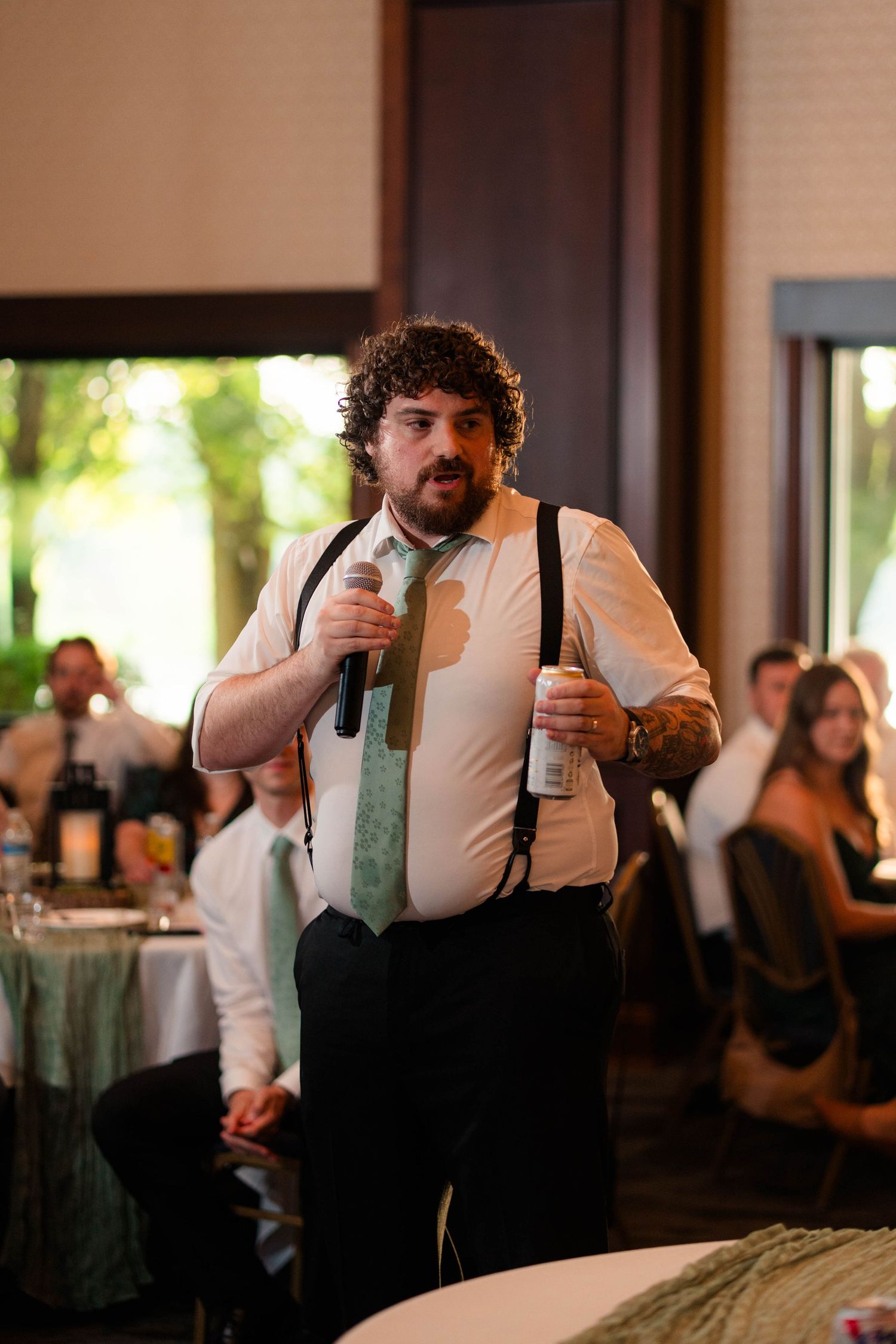 Curly-haired groomsman in suspenders and green tie delivers speech with microphone at wedding reception.