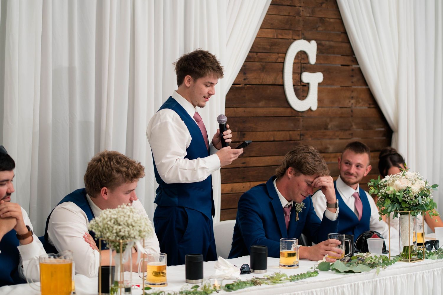 Young groomsman delivers a heartfelt speech at a wedding reception while guests laugh at the head table.