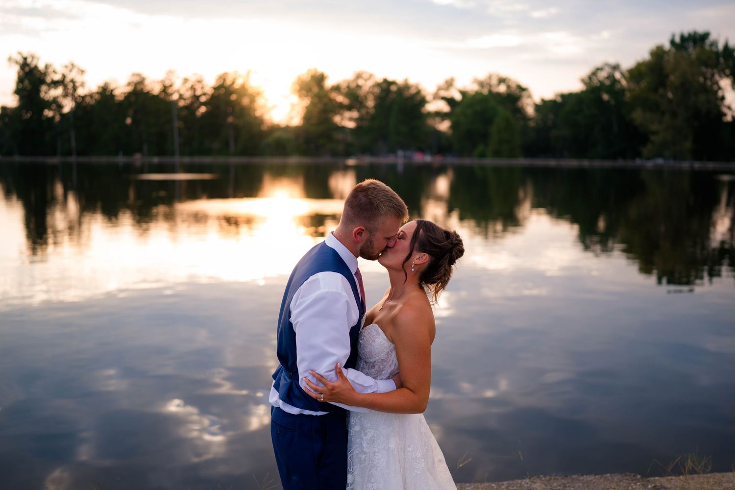 Bride and groom share a romantic kiss by a serene lake at golden sunset, surrounded by lush trees.