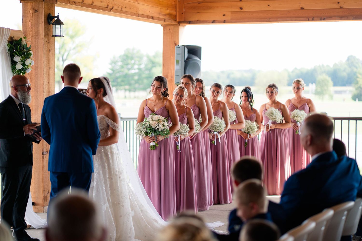 Bridesmaids in matching mauve dresses stand beside the bride during an outdoor pavilion wedding ceremony.