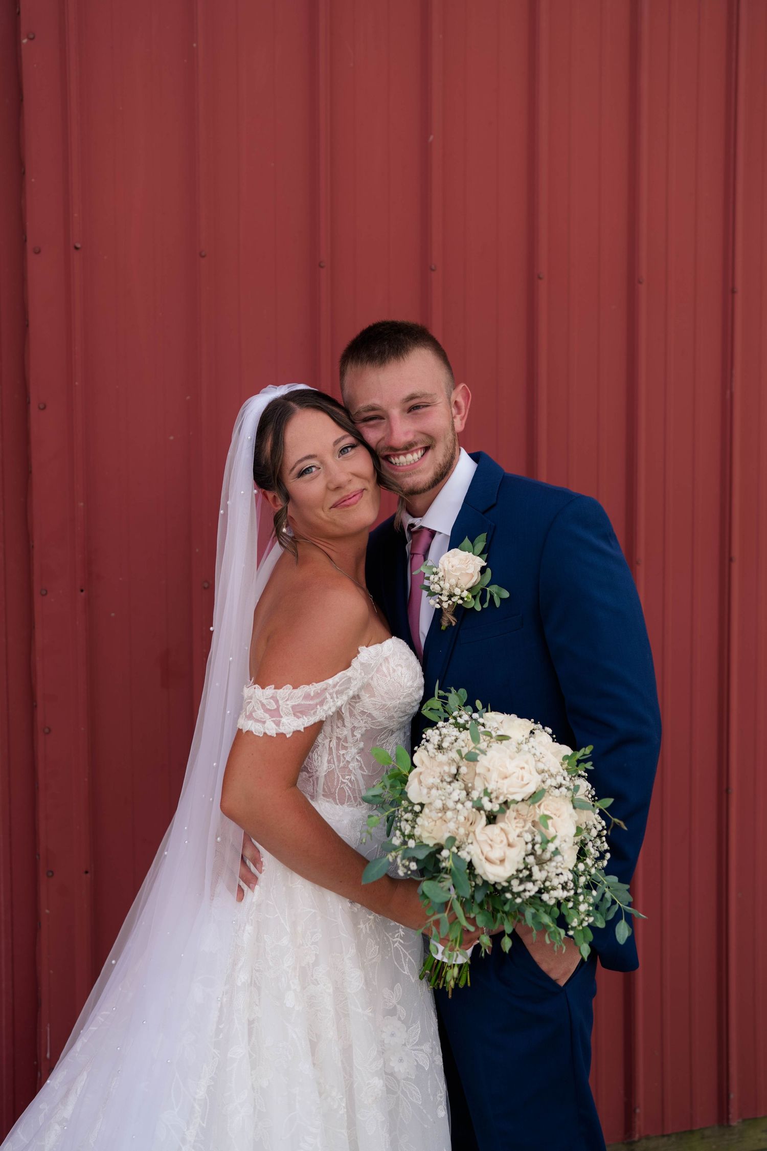 Happy bride and groom posing together against red barn wall, bride holding white rose bouquet.