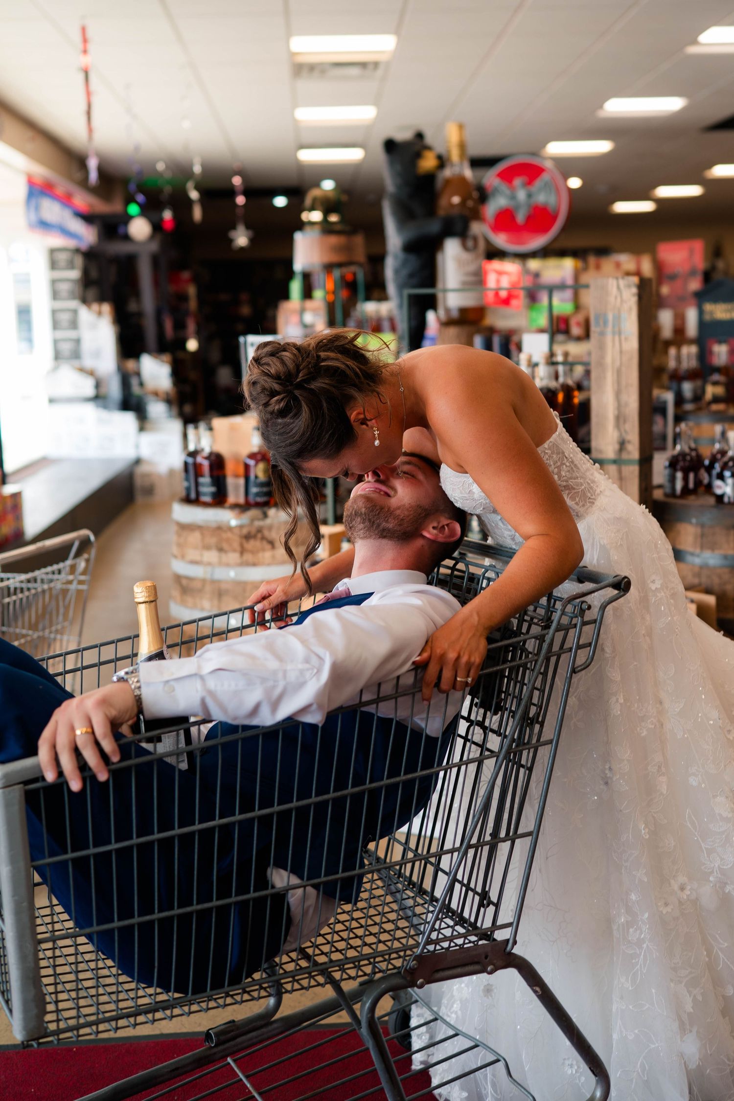 Bride leans over groom lying in shopping cart with champagne in fun wedding photo.