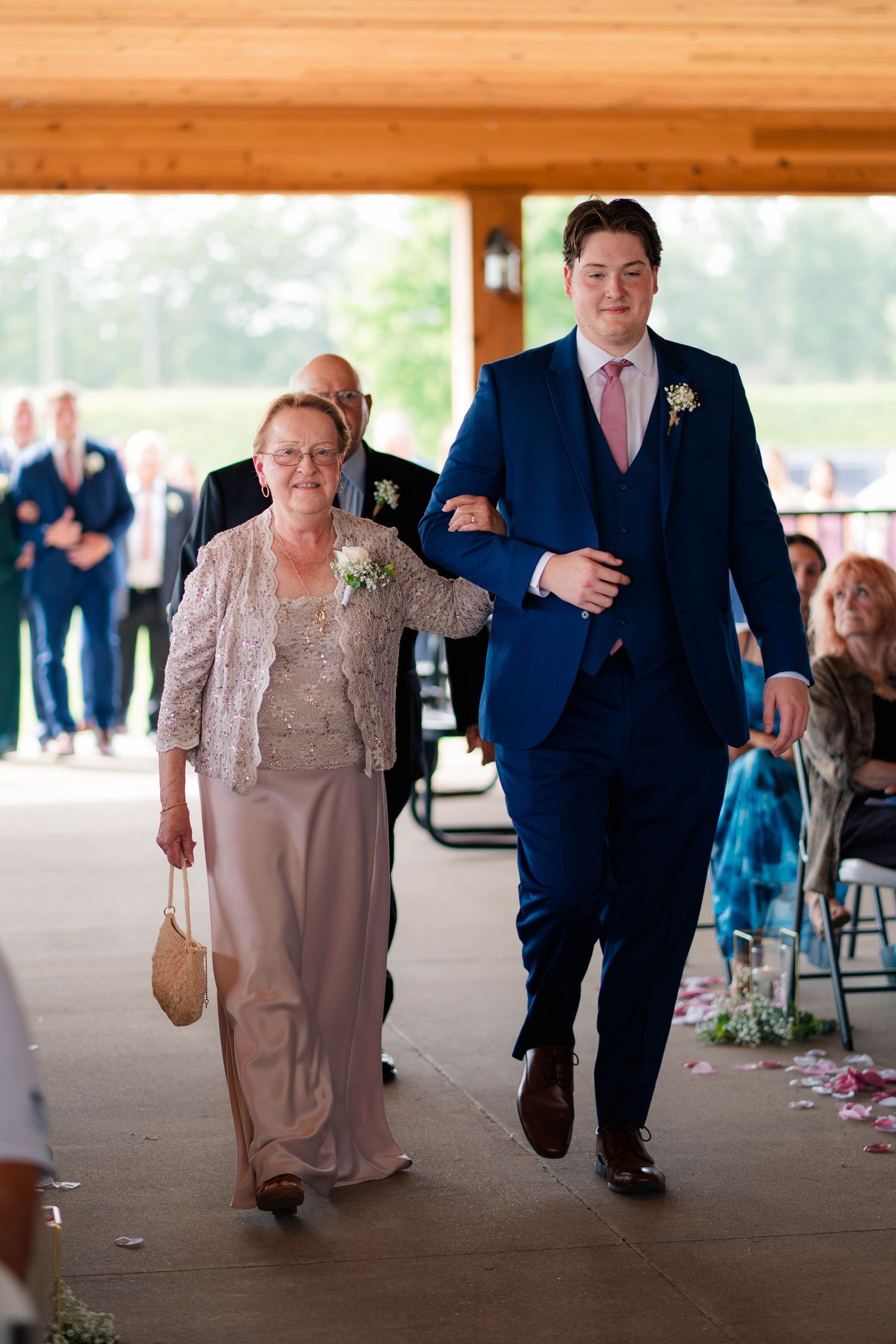 Groomsman in navy suit escorts elderly woman down wedding ceremony aisle.