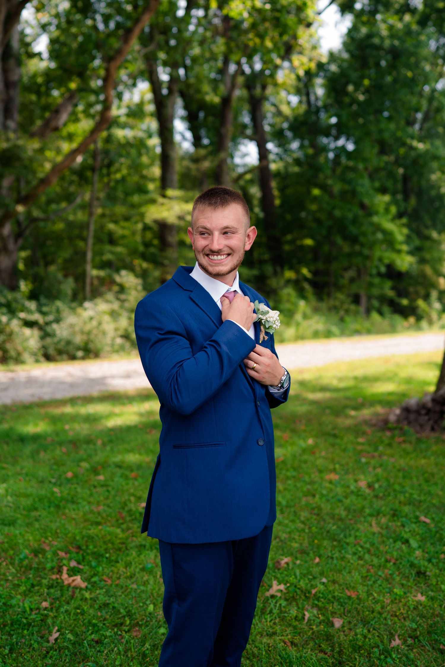 Smiling groom in navy suit adjusts boutonniere while standing on a green lawn outdoors.