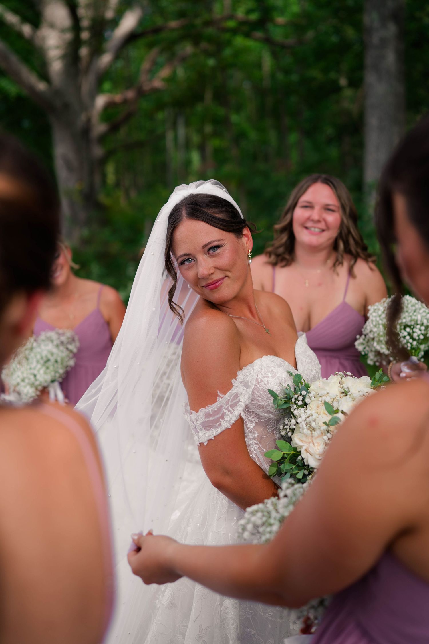 Smiling bride in lace off-shoulder gown and veil looks back at bridesmaids in purple dresses in forest setting.