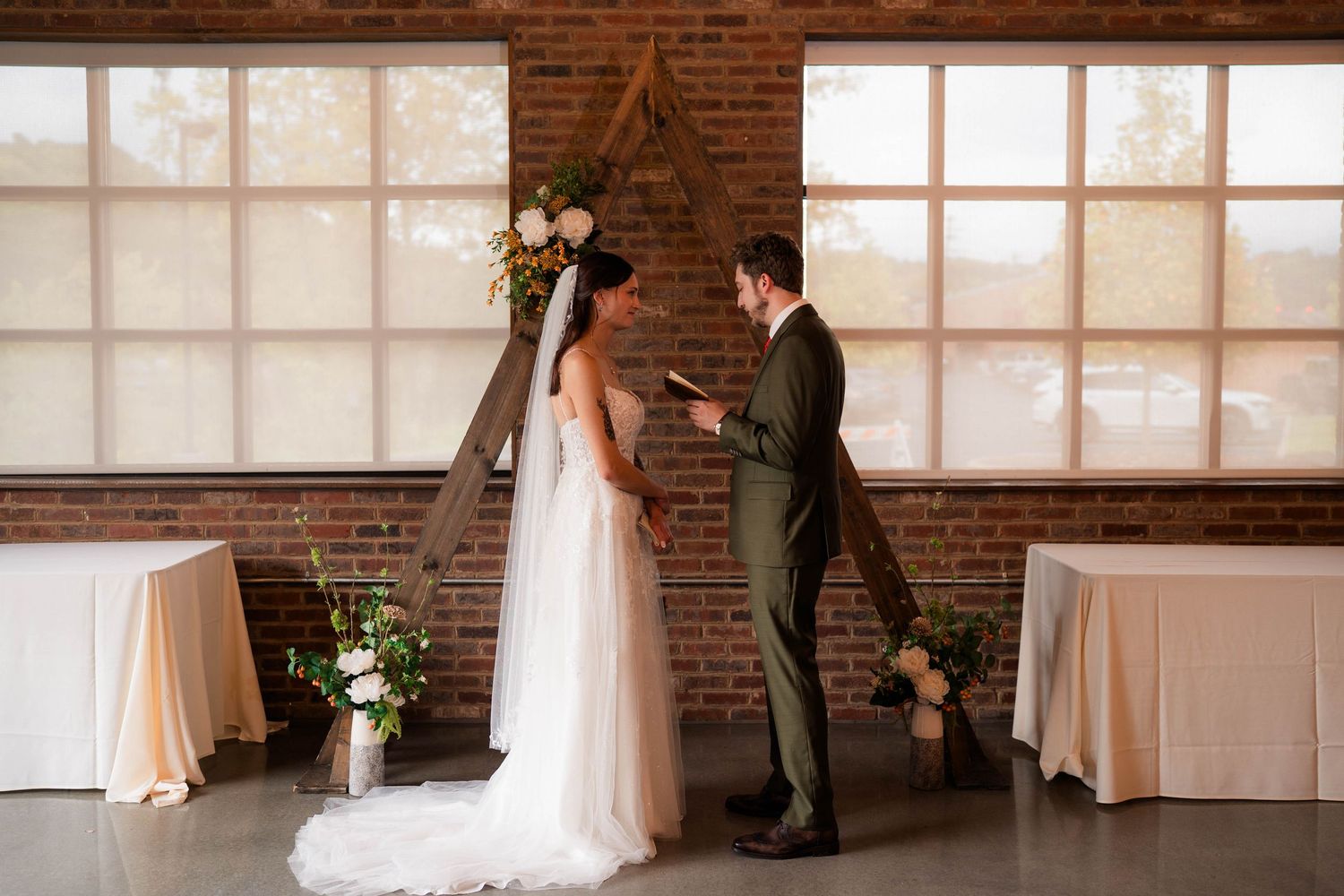 Bride and groom exchanging vows at rustic brick wedding venue with large windows and floral arrangements.
