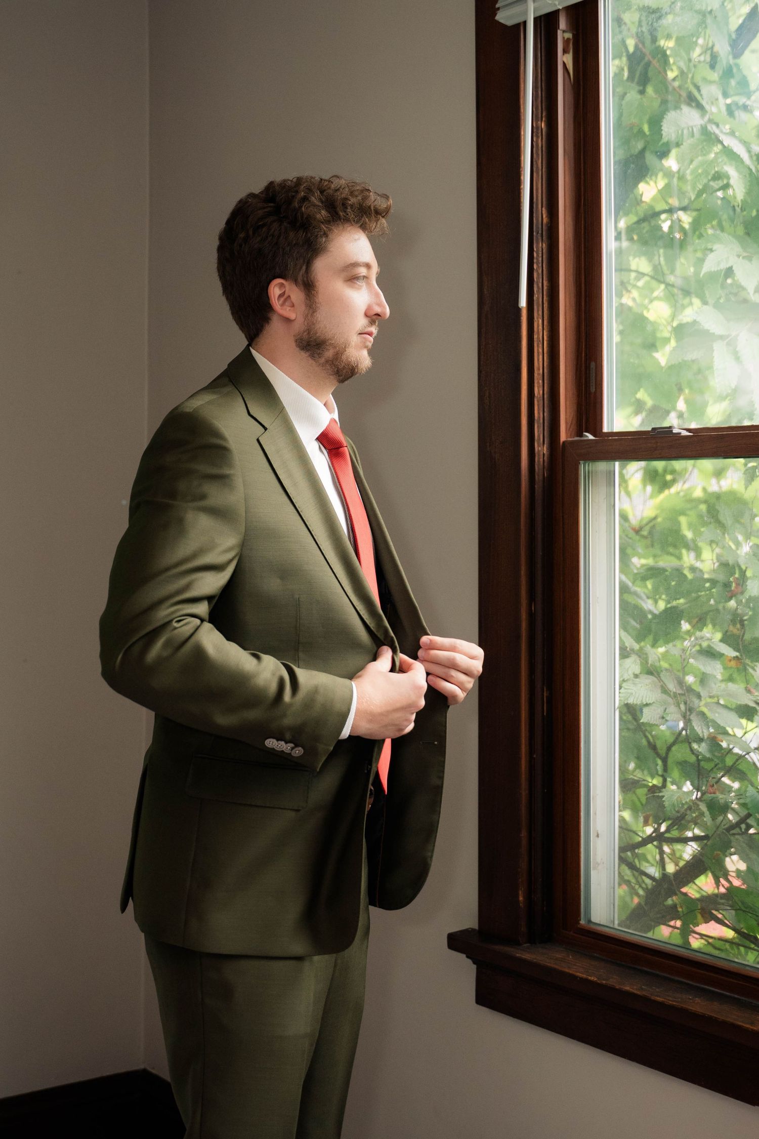 Groom in olive green suit and red tie gazing out window before wedding ceremony.