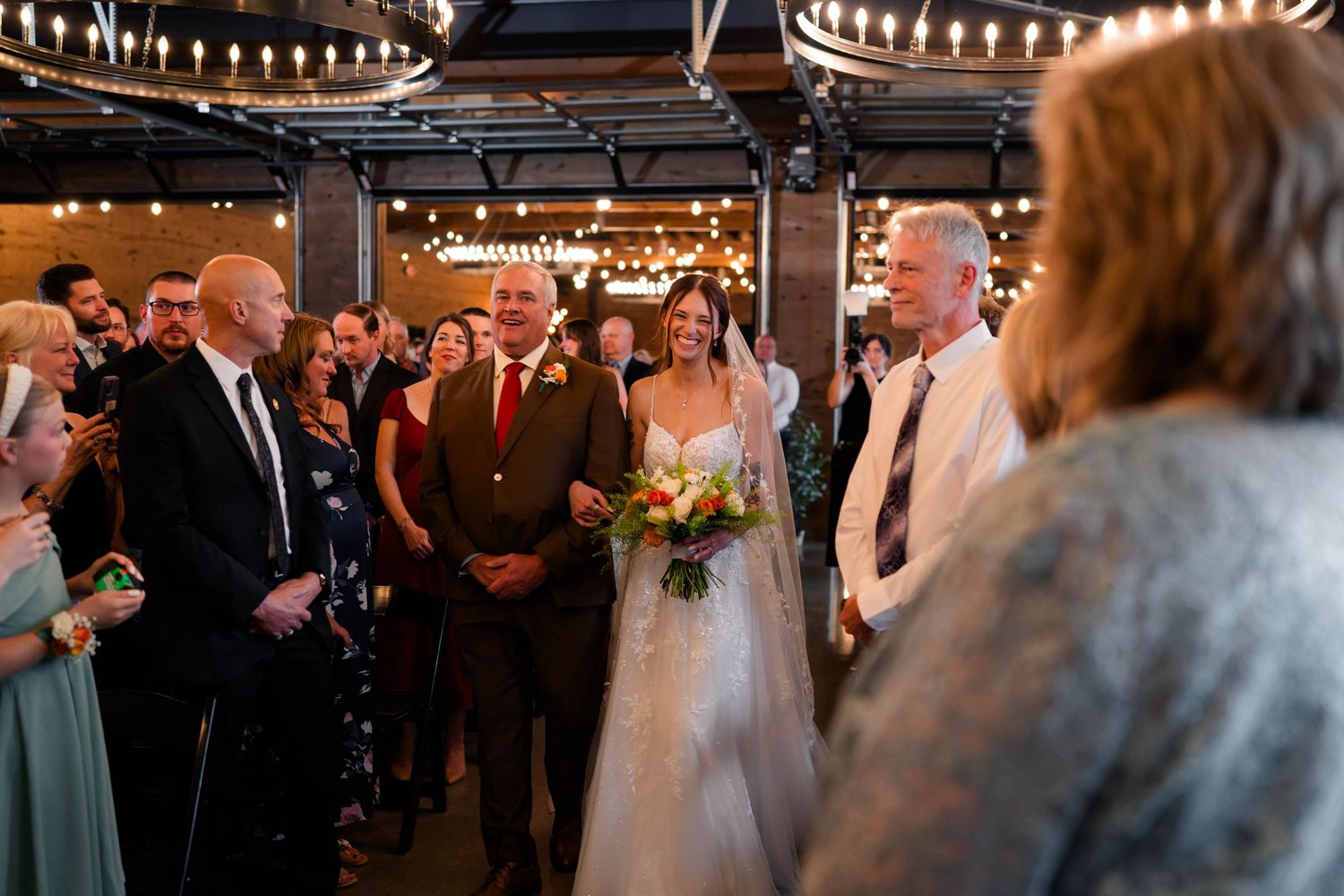 Bride carrying colorful bouquet walks down aisle with father at indoor wedding ceremony.