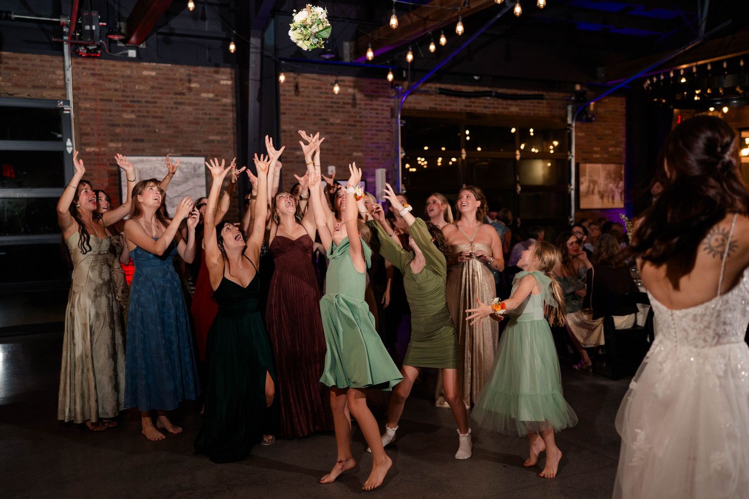Wedding guests reach up to catch bridal bouquet toss at indoor reception venue.