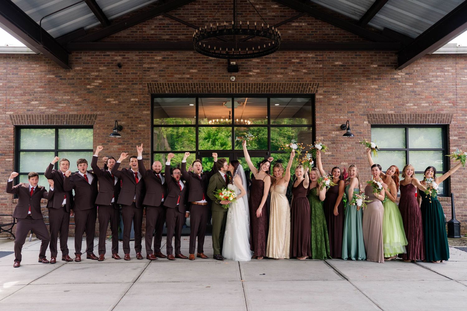 Large wedding party celebrates outside a brick venue, raising hands and bouquets in colorful attire.