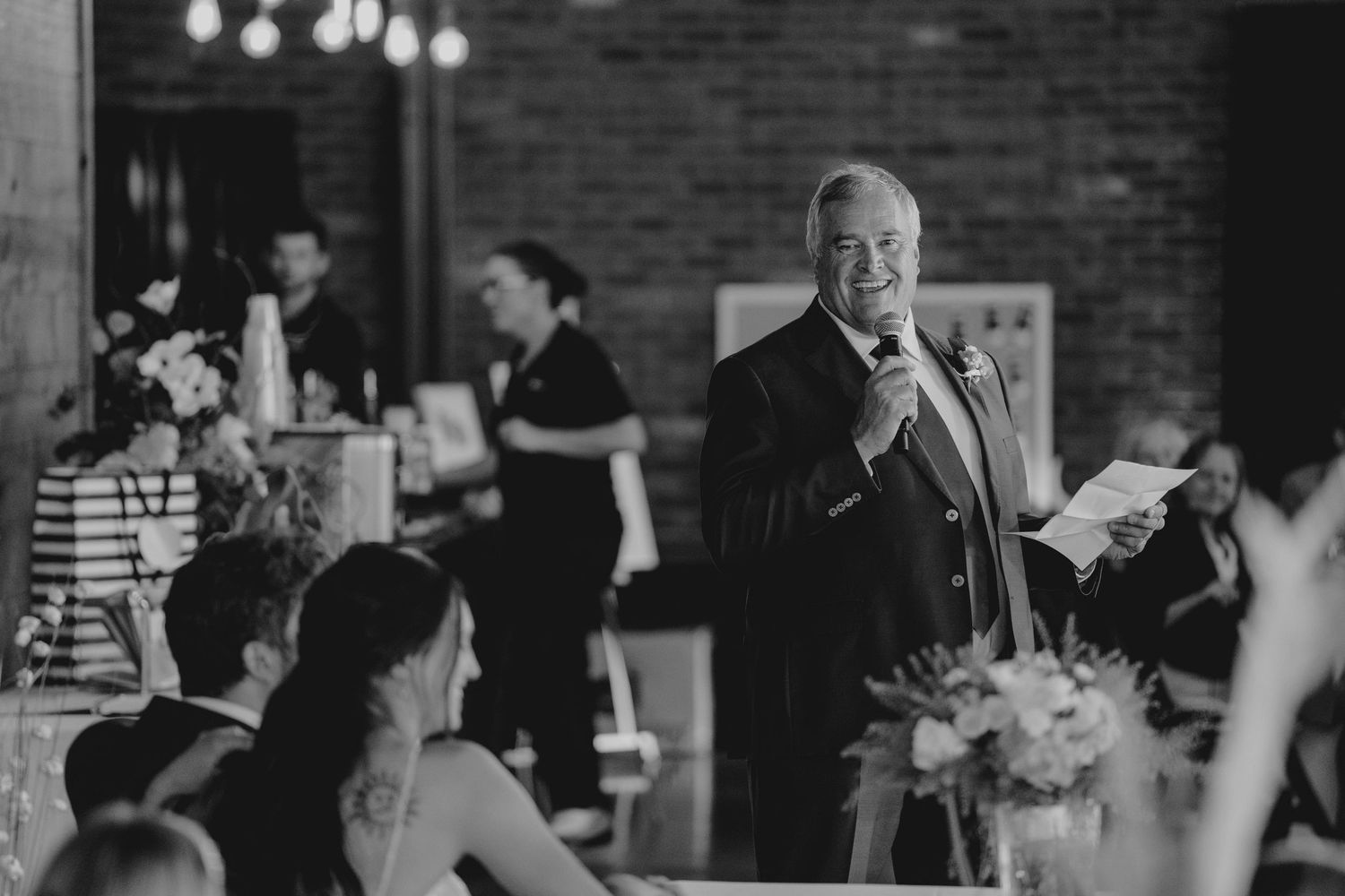 Man in suit smiling while giving a speech at a wedding reception in a rustic brick venue.