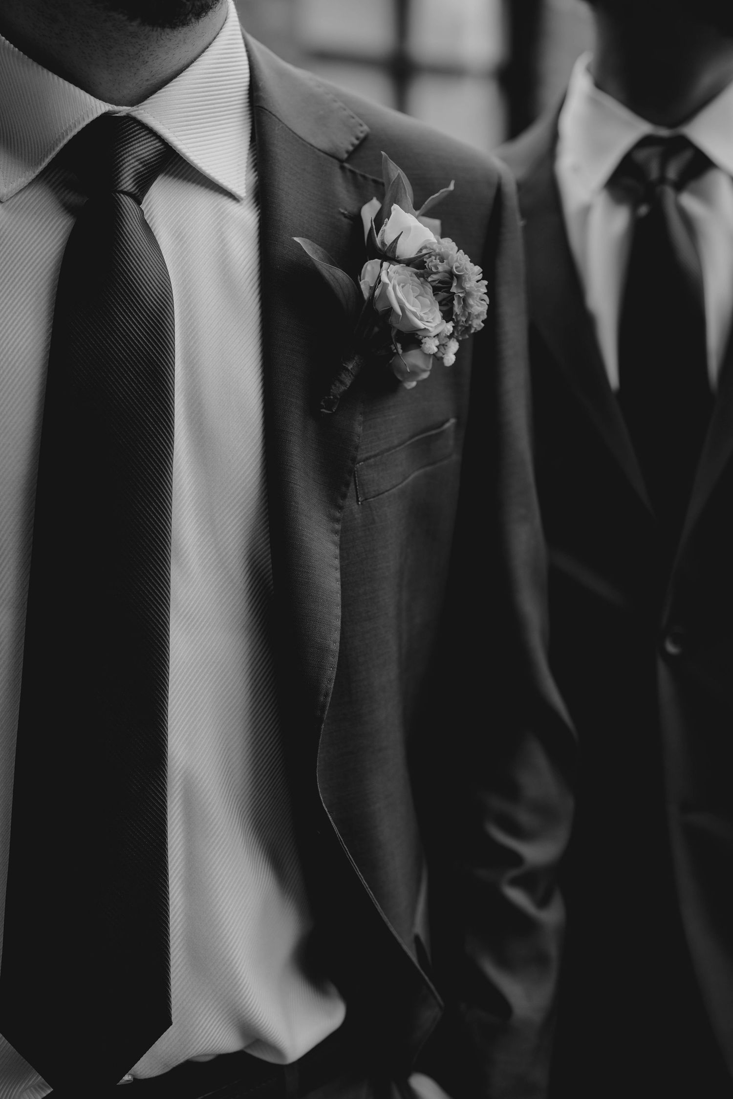 Black and white close-up of groom's boutonniere on a suit lapel with groomsmen standing nearby.