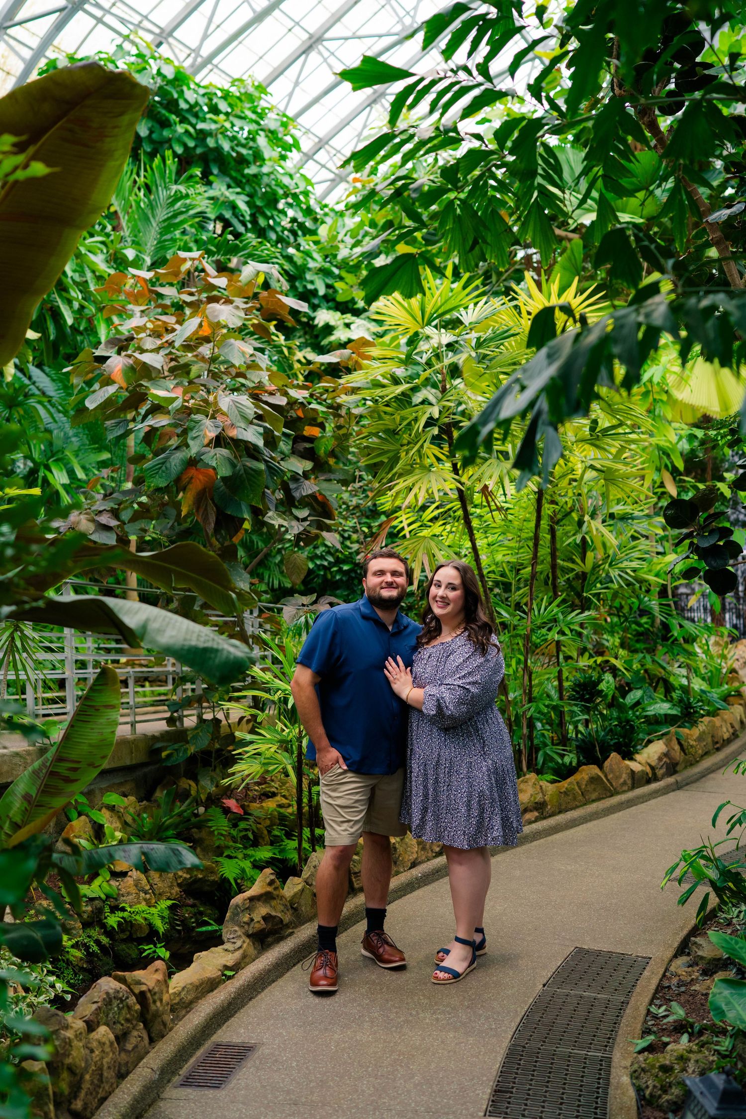 Smiling engaged couple standing together on a path in a stunning tropical greenhouse.