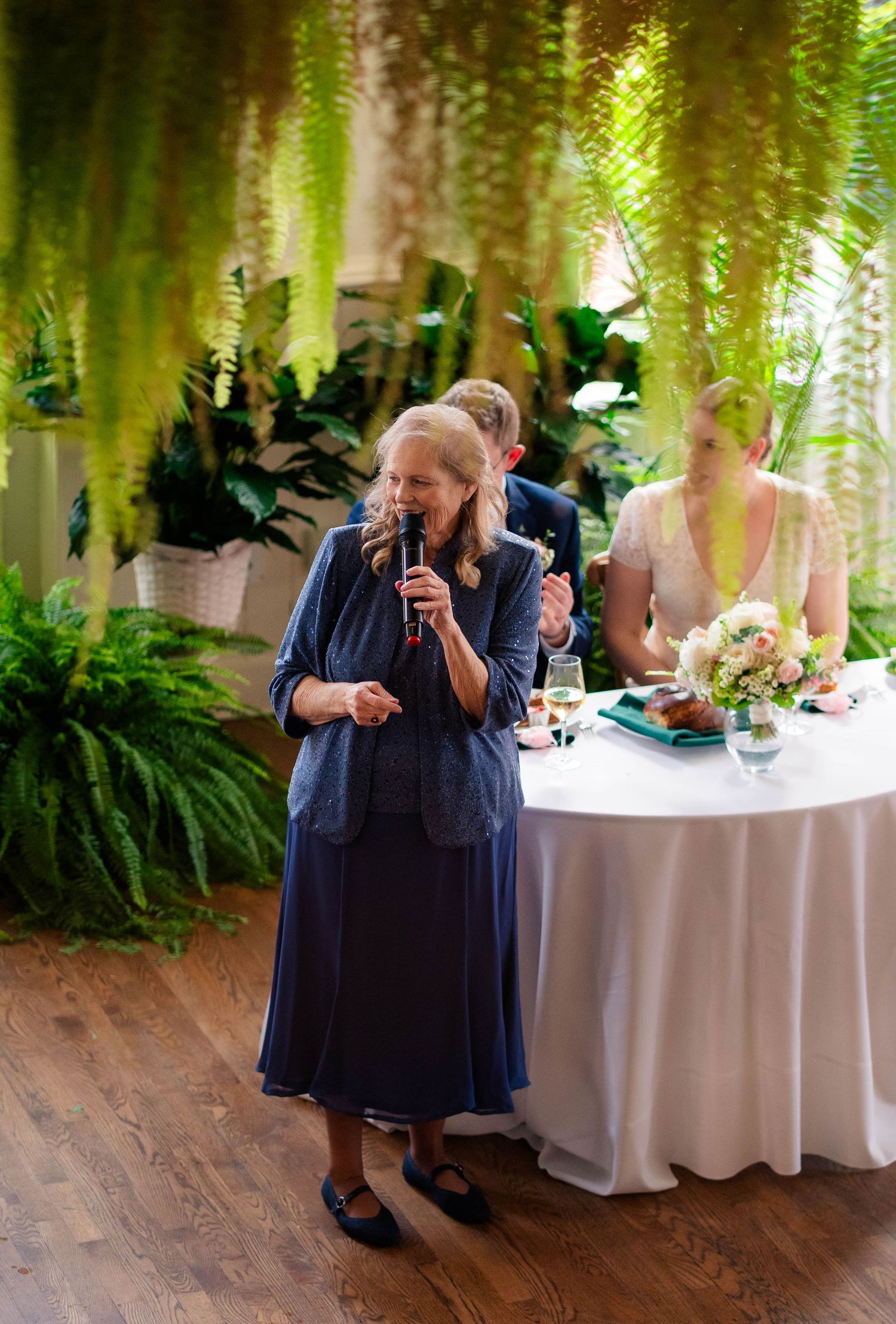 Elderly woman in blue dress giving a heartfelt speech at an elegant wedding reception with tropical decor.