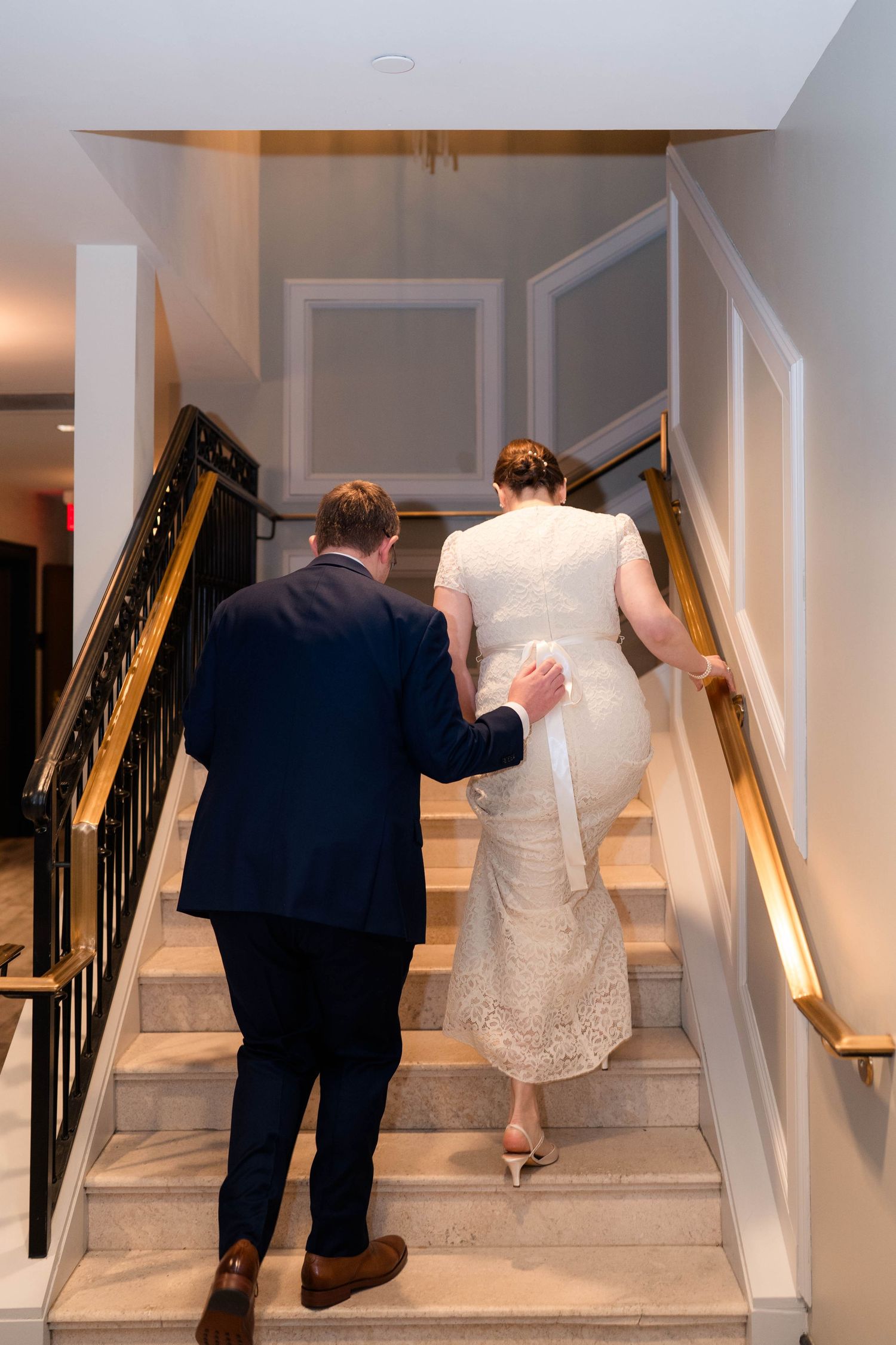 Bride in lace gown and groom in navy suit walk up elegant marble staircase together.