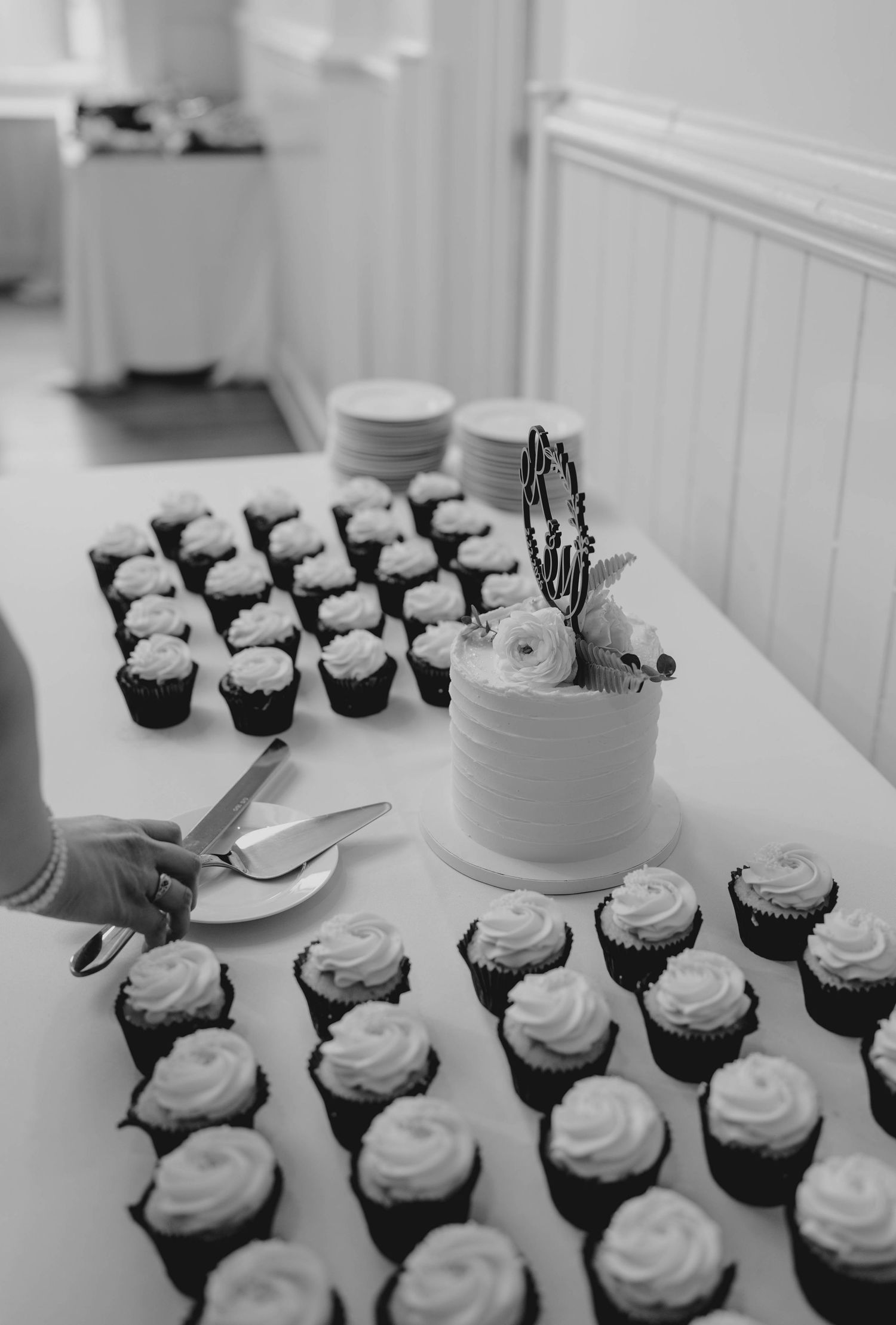 Black and white photo of wedding cake surrounded by chocolate cupcakes with white frosting on reception table.