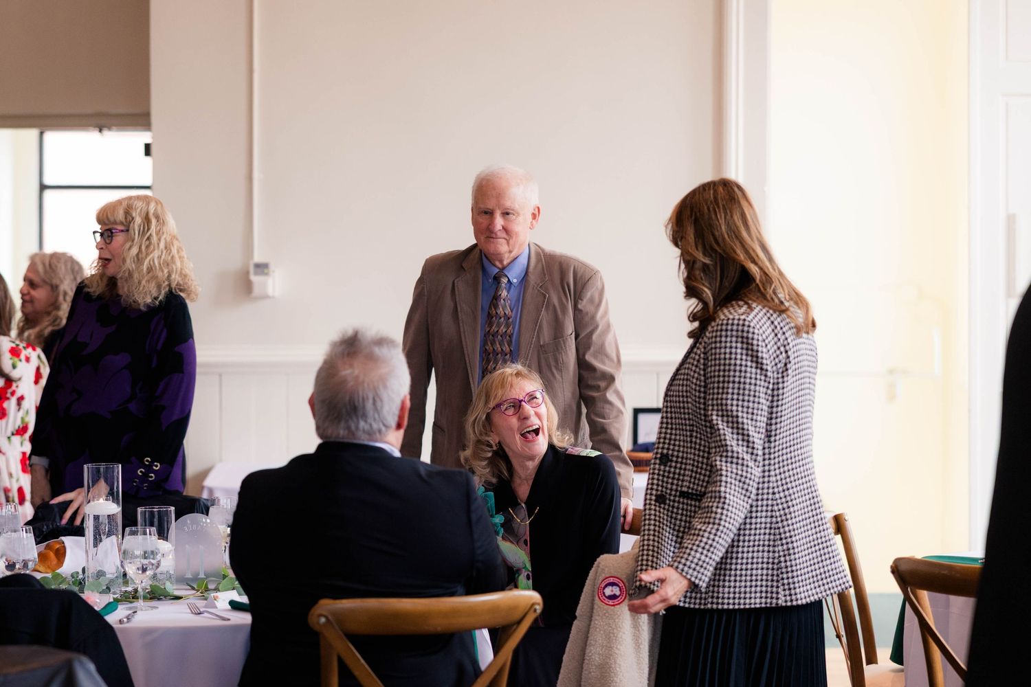 Guests engage in conversation at a formal luncheon event with white tablecloths and cross-back chairs.