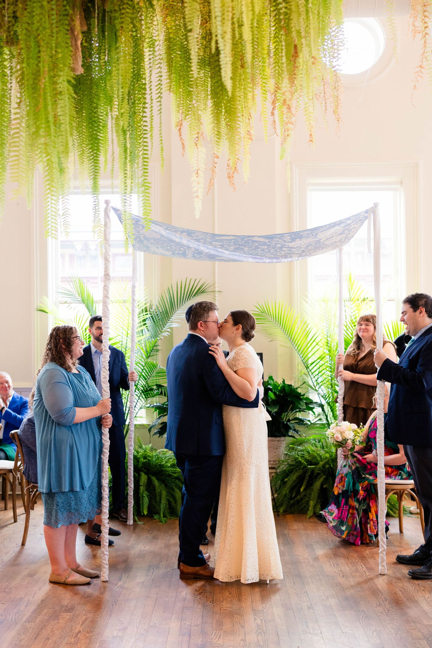 Newlyweds share first kiss under hanging fern chuppah surrounded by wedding party at indoor Jewish ceremony.