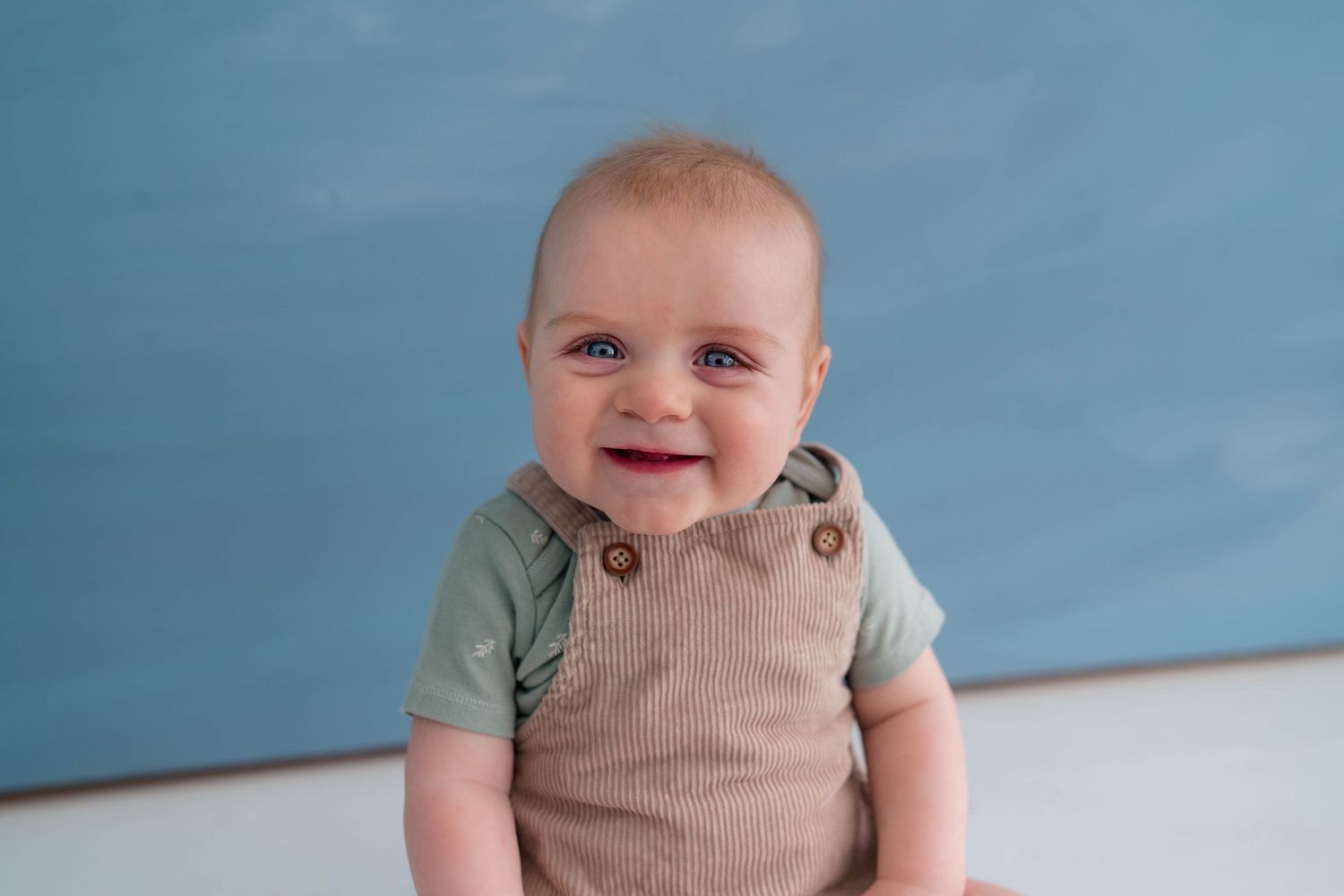 Smiling baby boy in tan overalls and green shirt sits against a blue background.