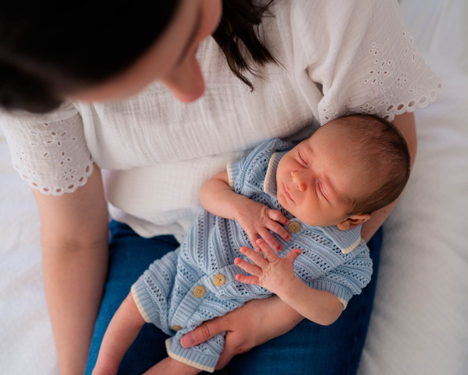 Mother in white top looking down lovingly at sleeping newborn baby dressed in a blue knit romper.